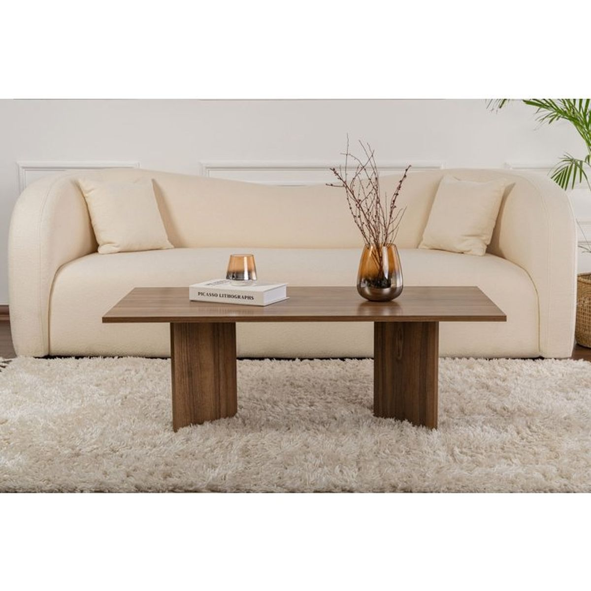 Ein Holztisch mit Büchern und einer Vase steht auf einem weißen Teppich vor einem cremefarbenen Sofa.