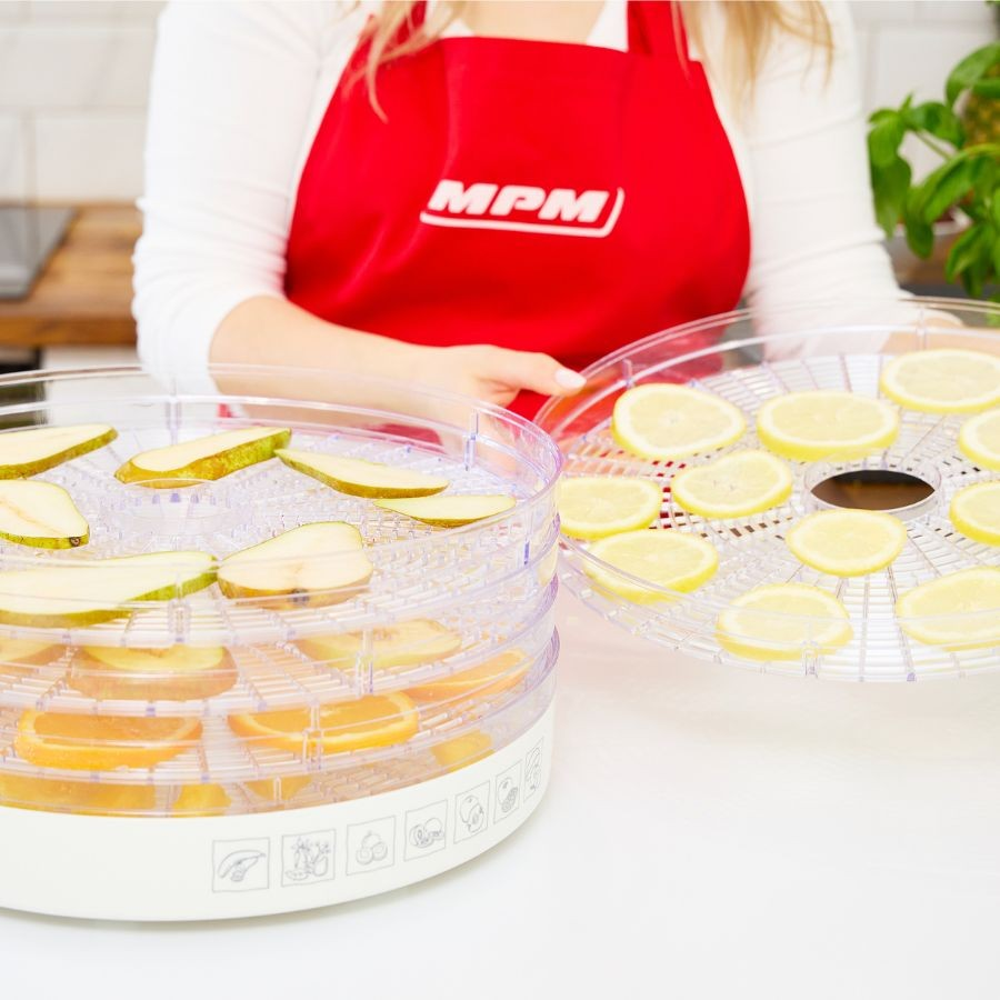 Una mujer con delantal rojo sostiene bandejas de deshidratador con peras, limones y naranjas.