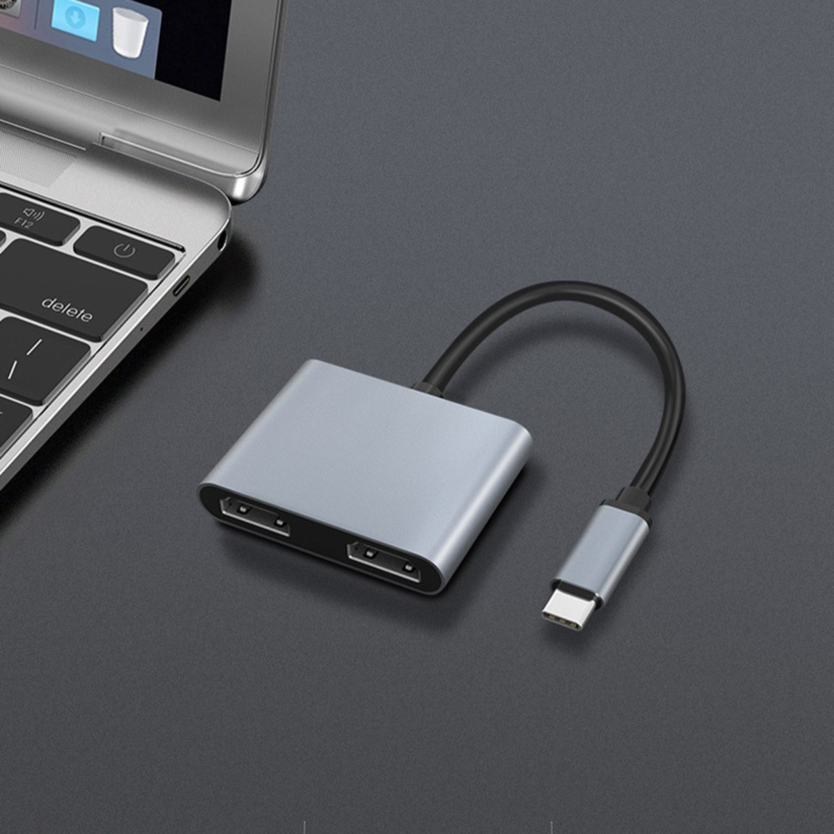 Srebrny hub USB-C z kablem leży obok laptopa na ciemnej powierzchni.