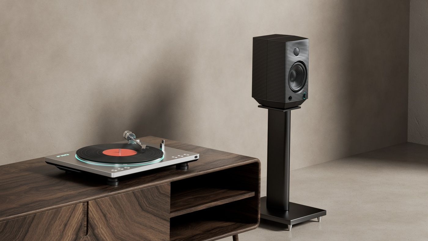 Gramofon na drewnianej szafce i czarny głośnik na stojaku przed szarą ścianą.