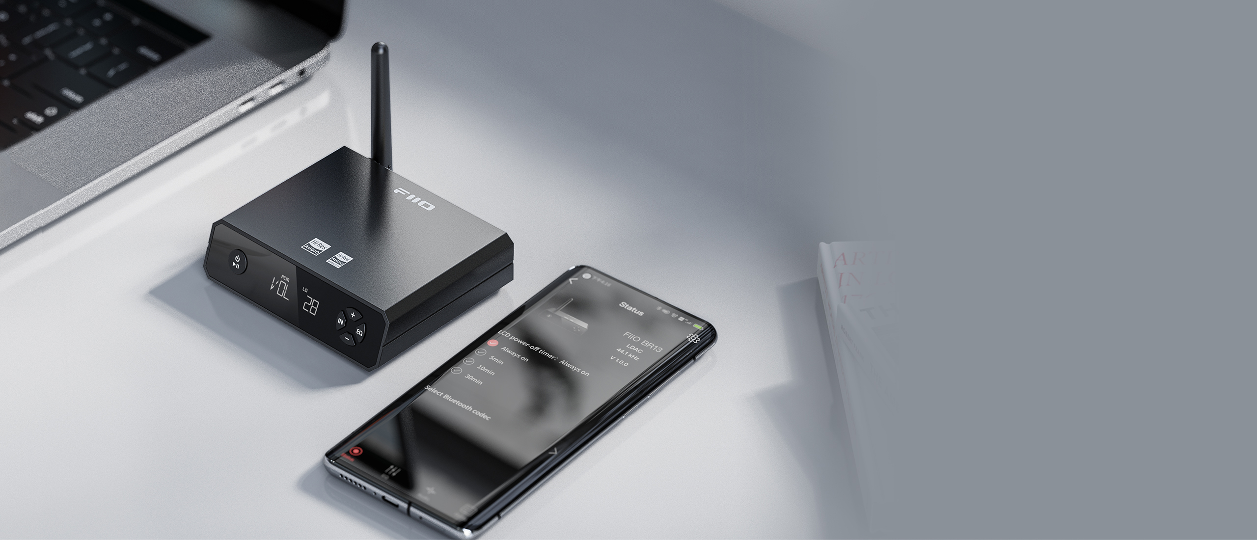Czarny wzmacniacz audio FiiO z anteną i smartfon leżą na białym biurku.