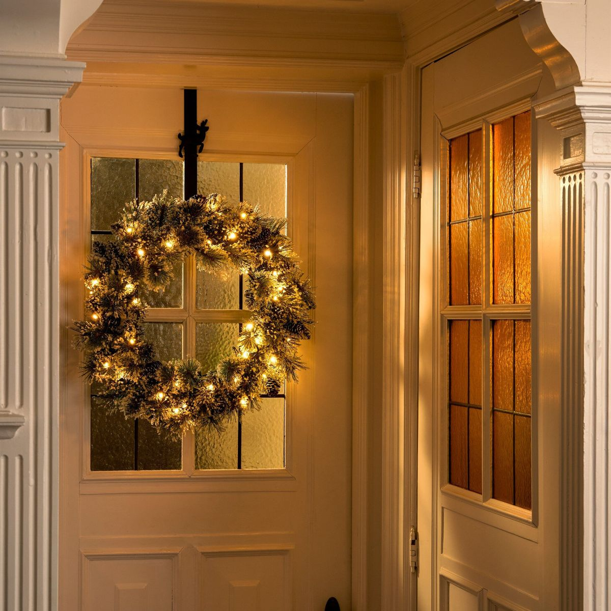 Een oplichtende kerstkrans met dennenappels hangt aan een witte glazen deur.
