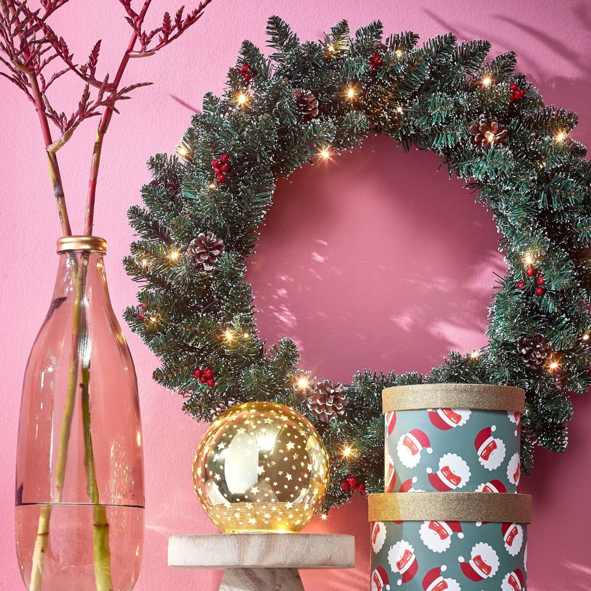 Een kerstkrans met lampjes, dennenappels en cadeauzen voor een roze muur.