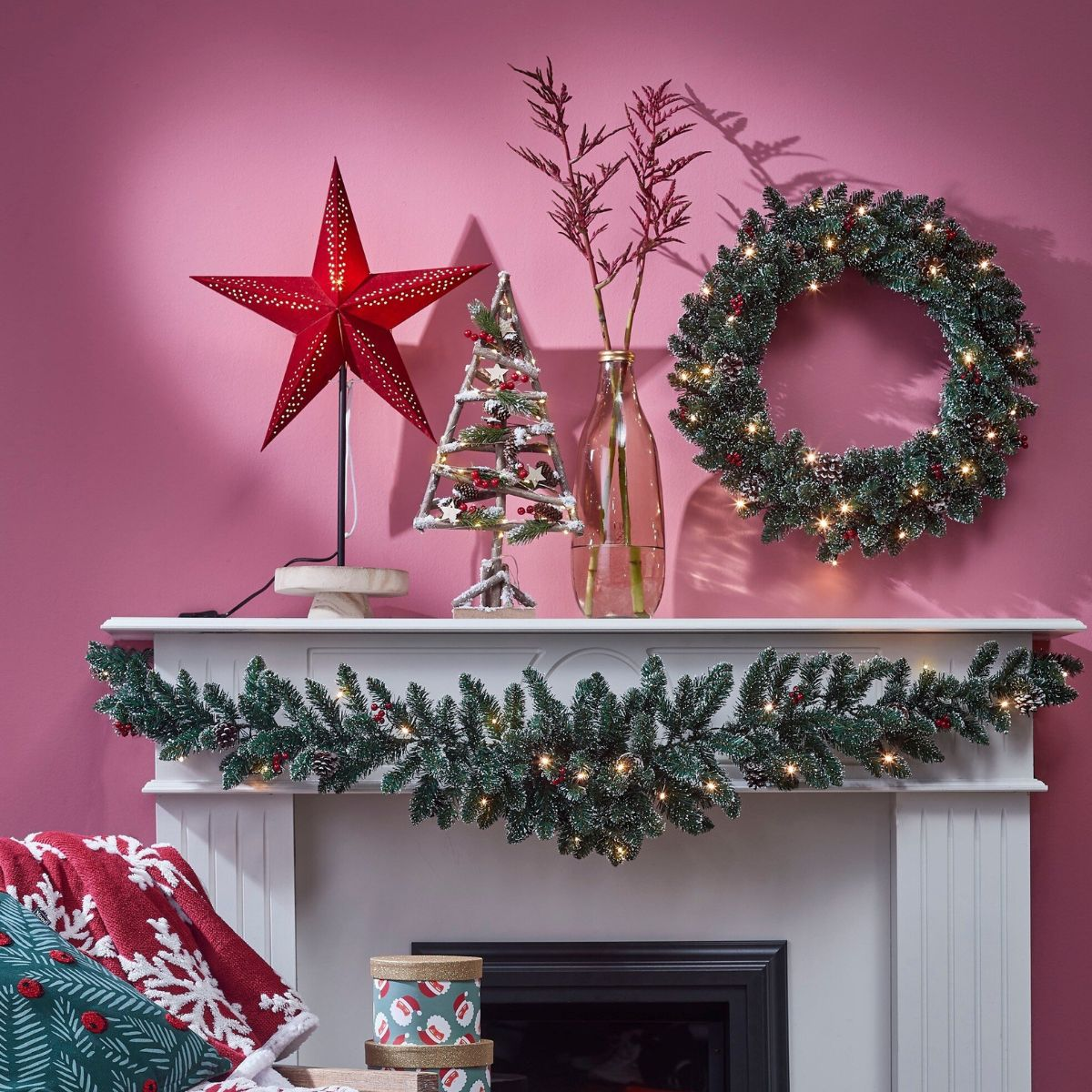 Kerstmanteldecoratie met rode ster, boompje, verlichte krans en slingers tegen een roze muur.