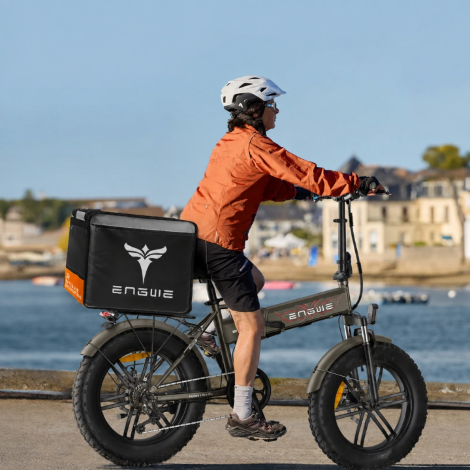 Una persona con casco bianco e giacca arancione guida una e-bike vicino al mare.