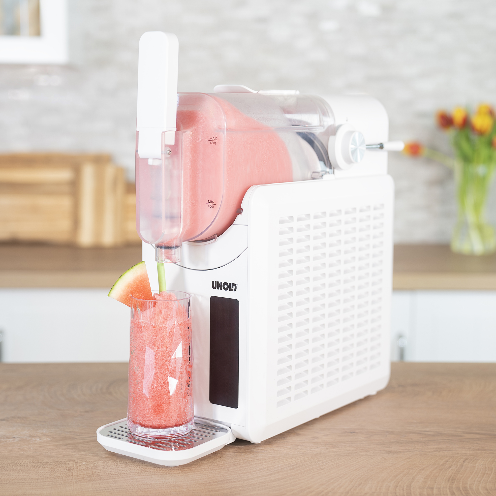 Eine weiße Unold Slush-Maschine auf einer Holztheke füllt pinkes Slush in ein Glas mit Wassermelone.