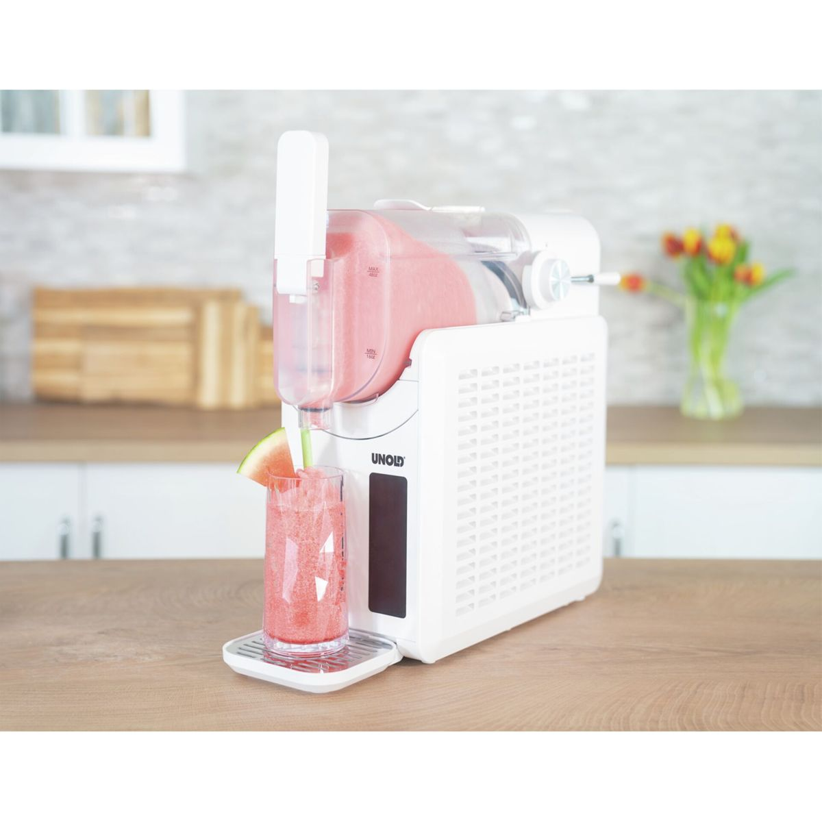 Eine weiße Slush-Eis-Maschine auf einem Holztisch füllt ein Glas mit rotem Getränk, garniert mit Wassermelone.