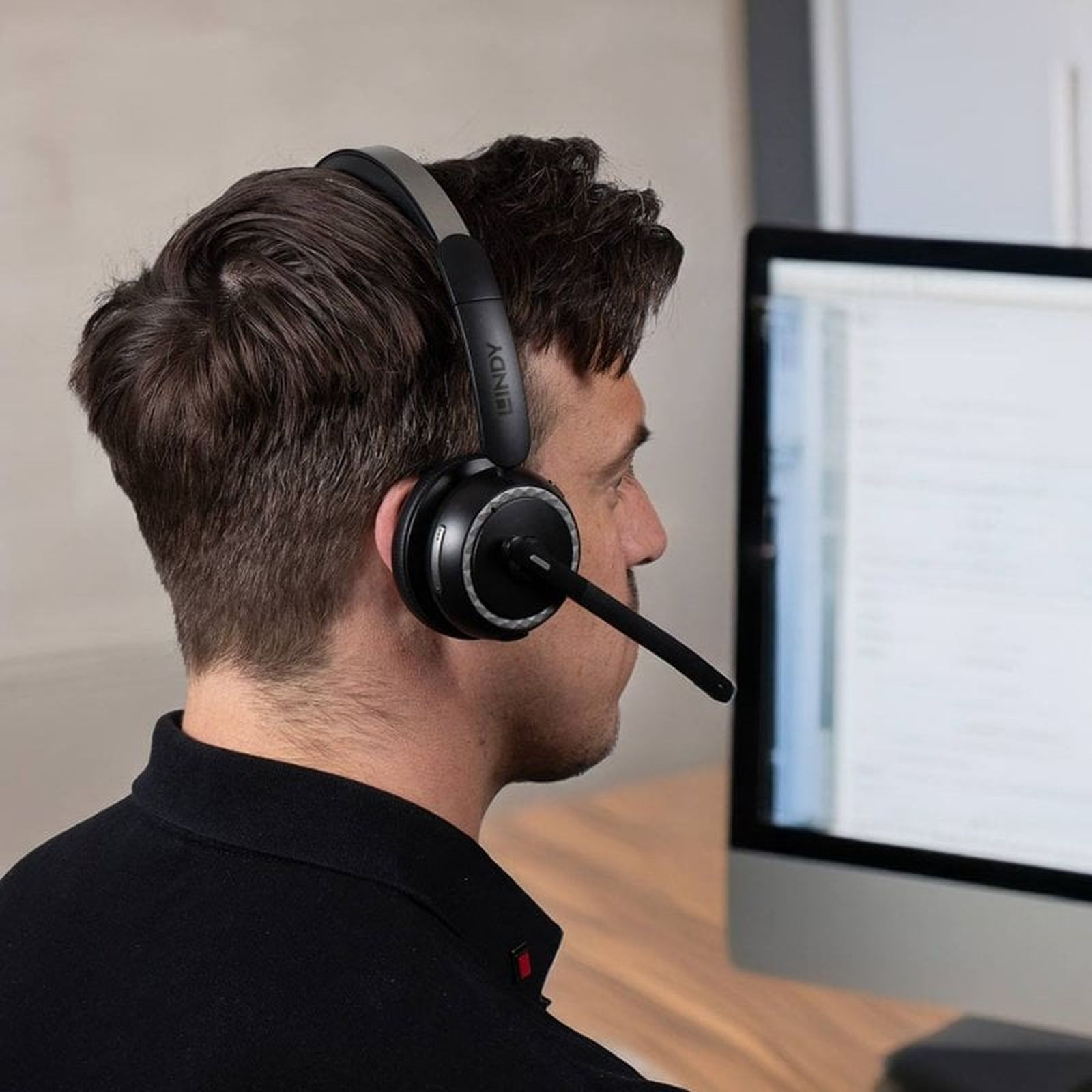Un hombre con camisa negra se sienta frente al monitor de una computadora, usando auriculares con micrófono.