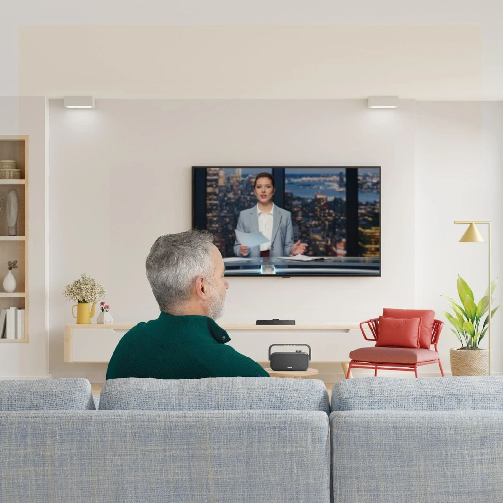 Siwiejący mężczyzna siedzi na niebieskiej kanapie, oglądając telewizję w jasnym salonie.
