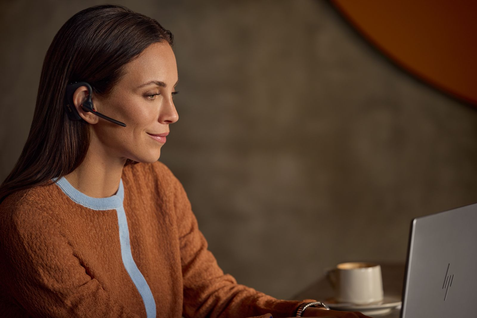 Una mujer con suéter naranja trabaja en una computadora portátil usando auriculares.
