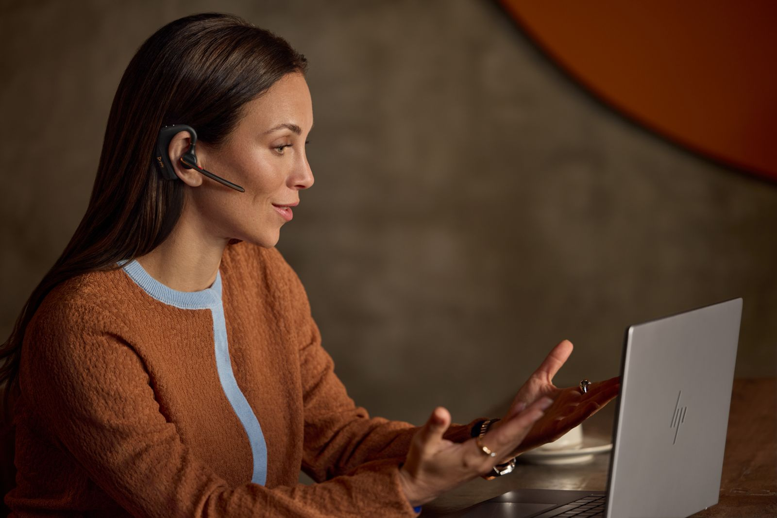 Una mujer con un jersey naranja y azul y auriculares está sentada frente a una laptop.