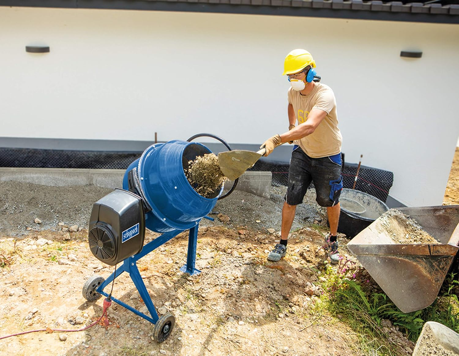 Un operaio con elmetto giallo pala del materiale in una betoniera blu.