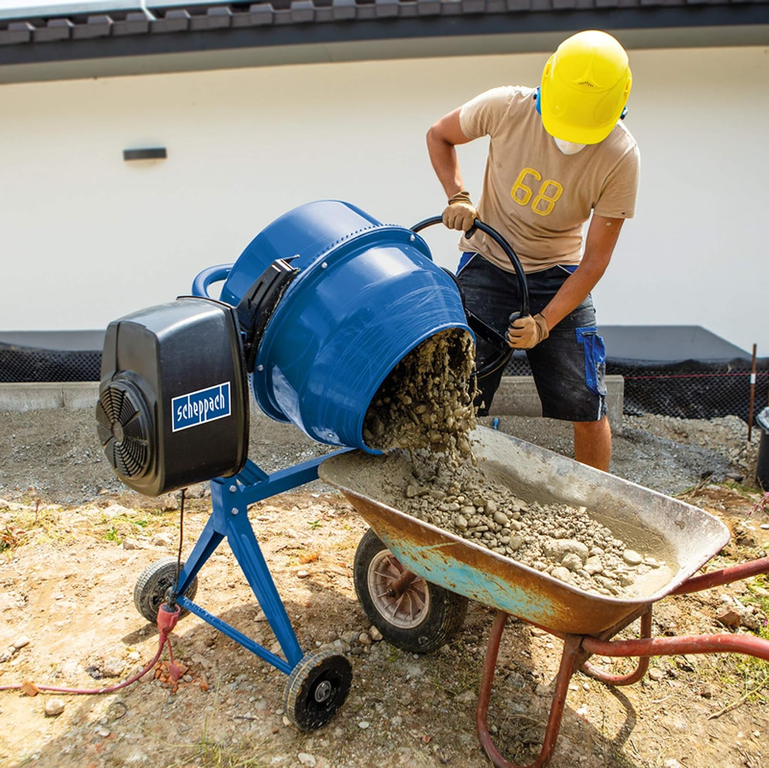 Un operaio con elmetto giallo versa calcestruzzo fresco da una betoniera blu Scheppach in una carriola.