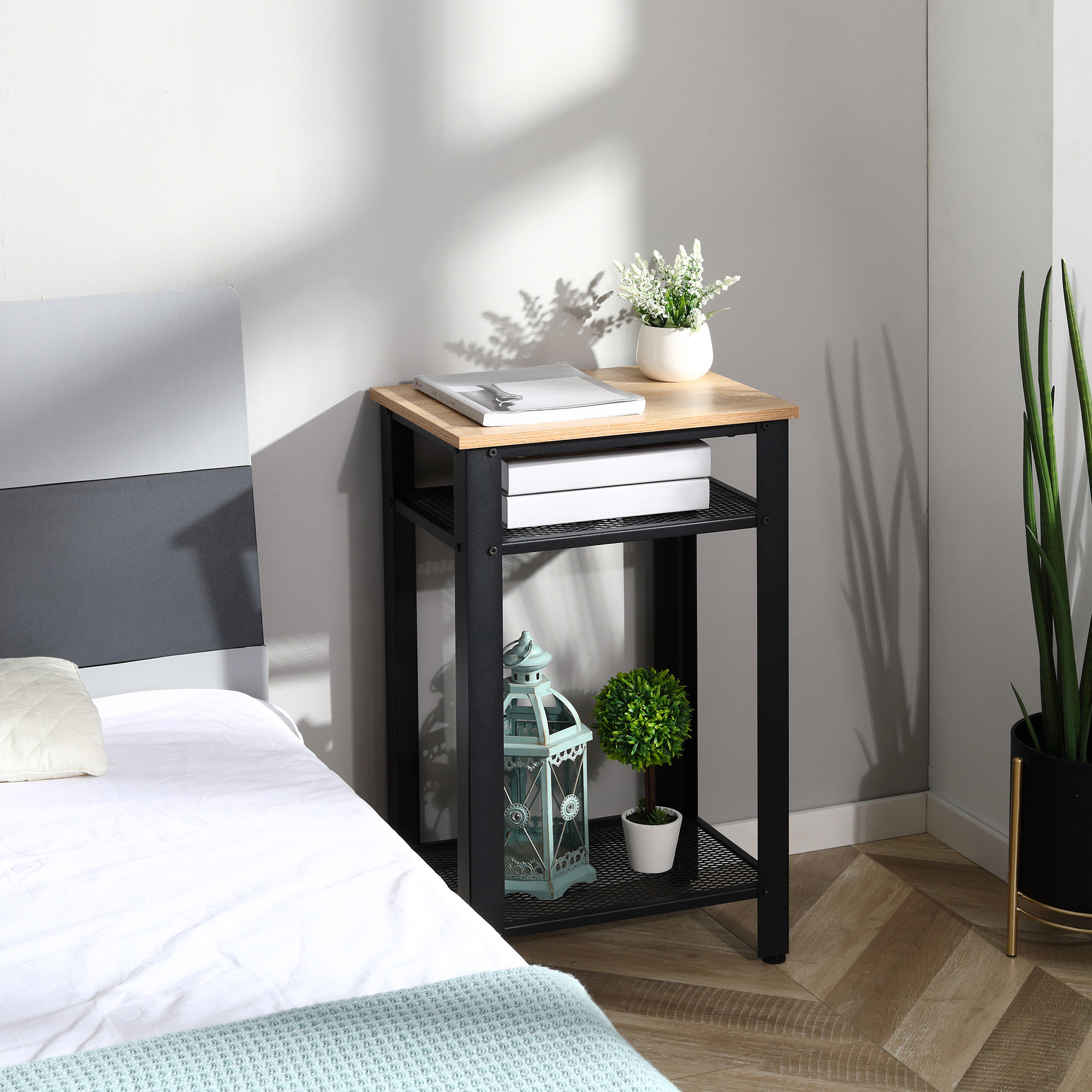 Una mesita de noche de madera y metal negro junto a una cama, con una planta, libros, un farol y decoración.