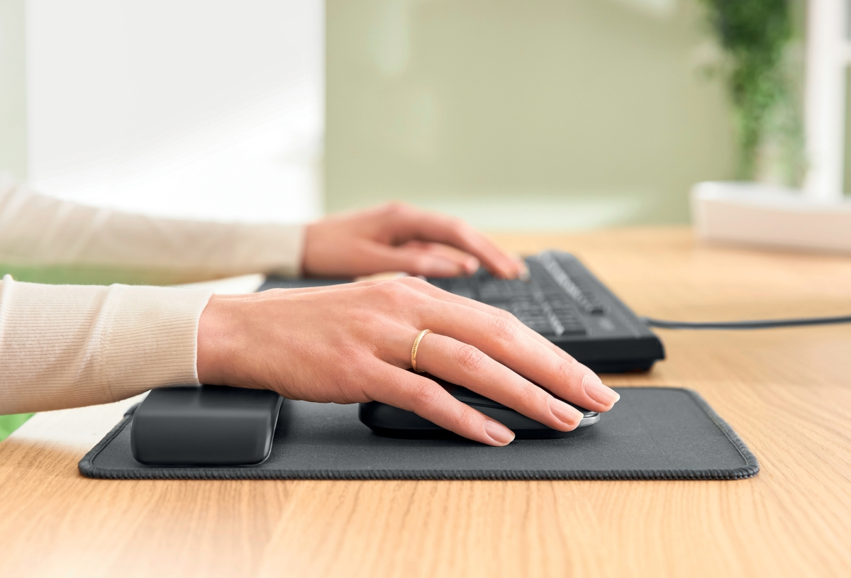 A hand wearing a ring uses a computer mouse on a wrist rest pad, with a keyboard in the background.