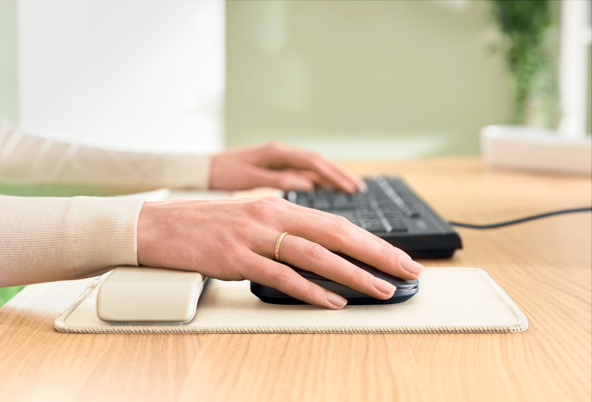 A person uses a computer mouse and keyboard on a wooden desk, resting their wrist on a support cushion.
