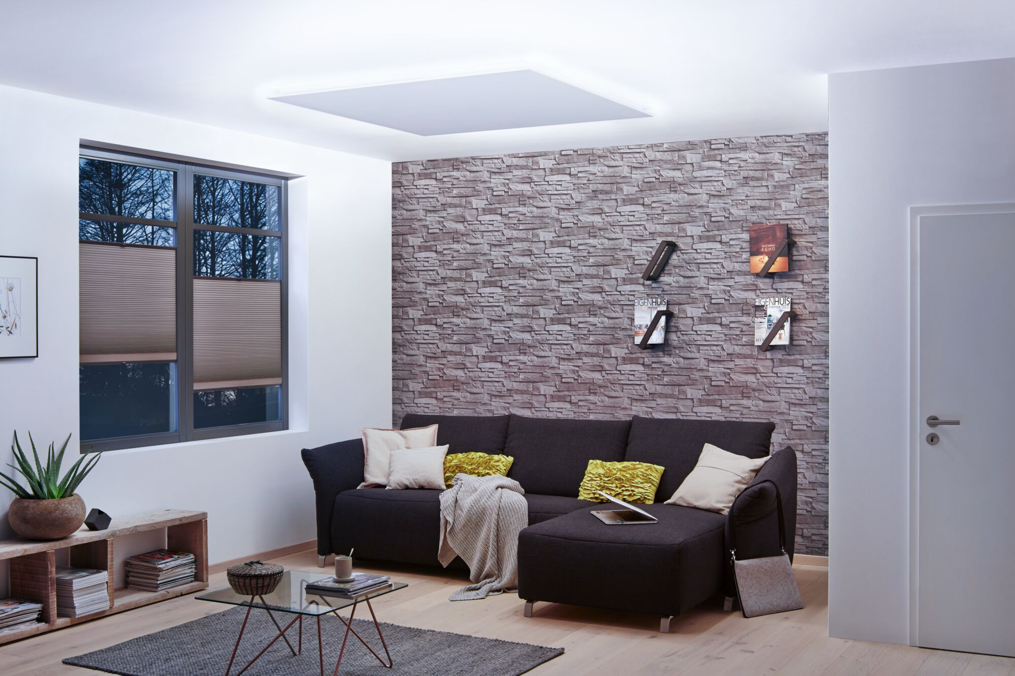 Moderne Wohnzimmer mit dunklem Sofa, Steinwand, Glastisch und großem Fenster mit Plissee-Rollos.