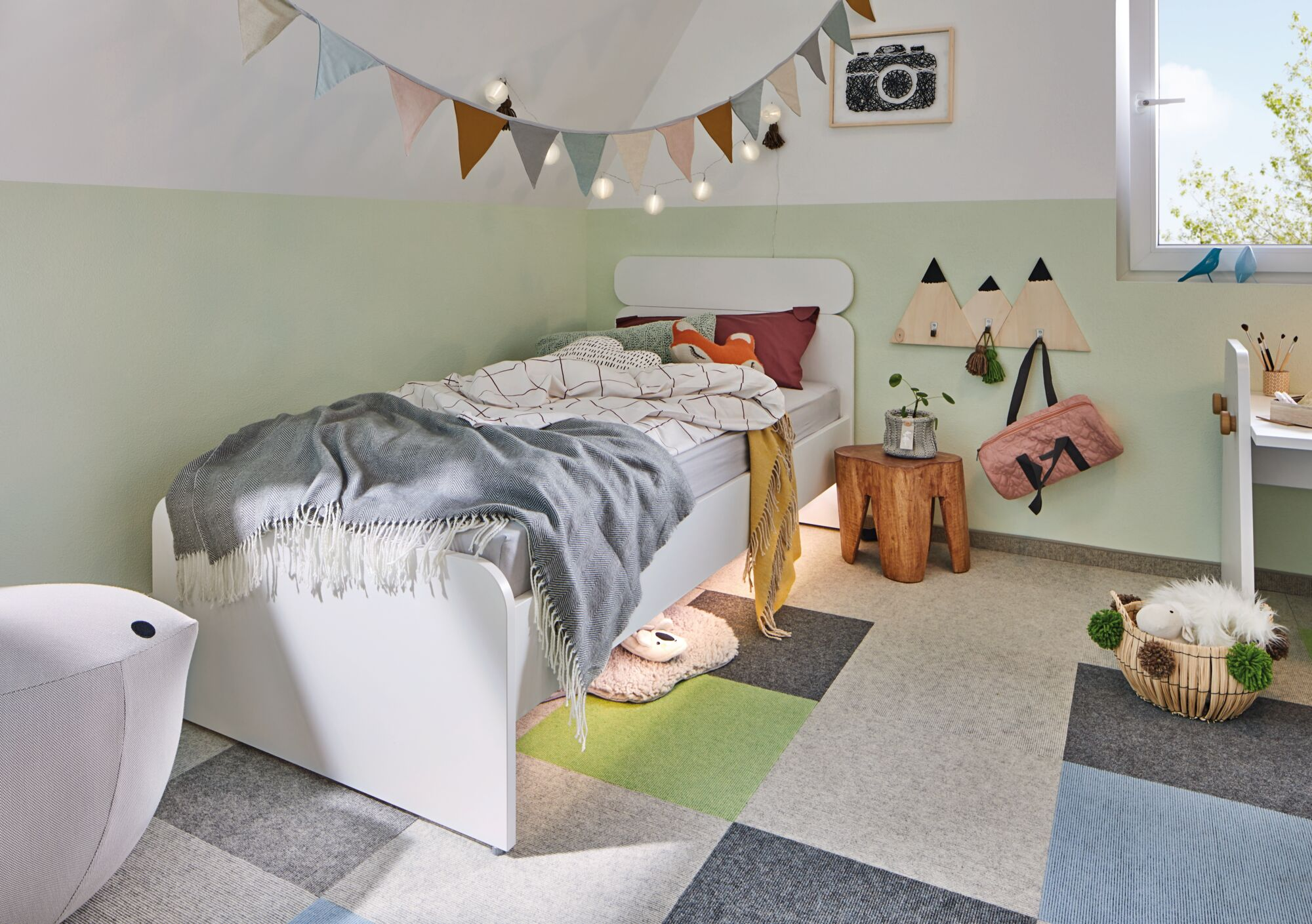 Gemütliches Kinderzimmer mit weißem Bett, grünen Wänden, bunten Wimpeln, Holztaburett und Spielzeugkorb.