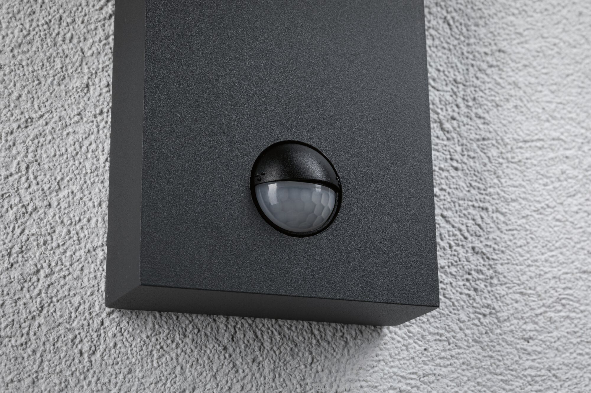 Ein grauer Wandkörper mit integriertem, rundem Bewegungsmelder auf einer weißen Putzwand.