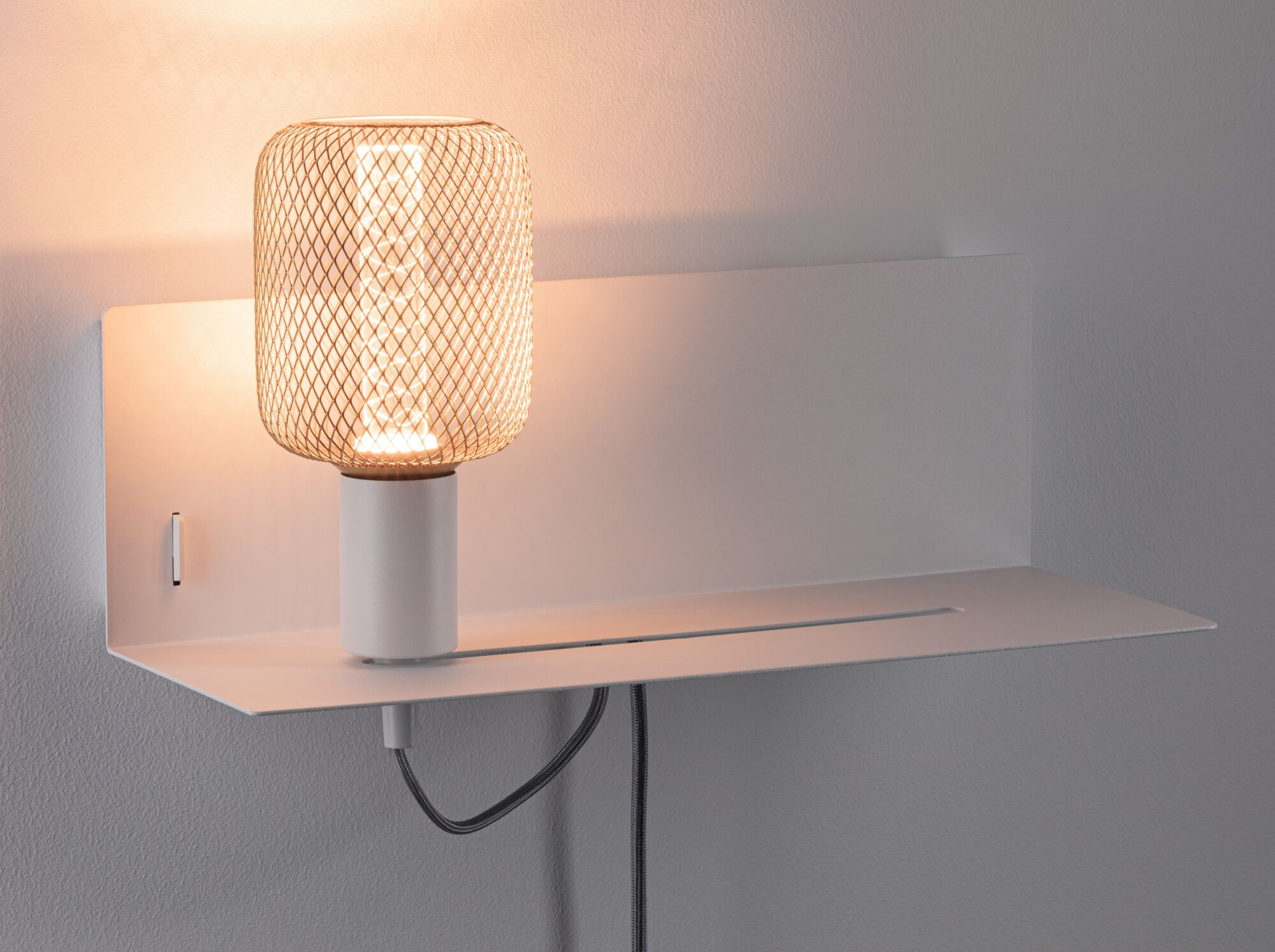 Eine leuchtende Tischlampe mit Metallgitter steht auf einem weißen Wandregal vor einer grauen Wand.