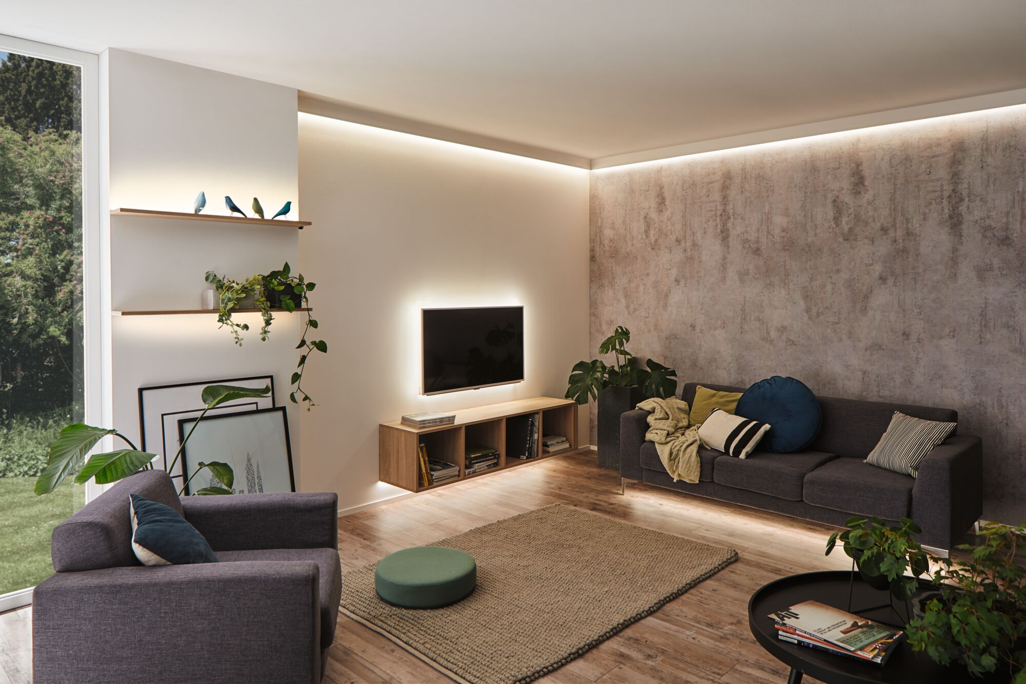 Wohnzimmer mit dunklem Sofa, Sessel, TV an der Wand, hängendem Regal mit Pflanzen und indirekter LED-Beleuchtung.