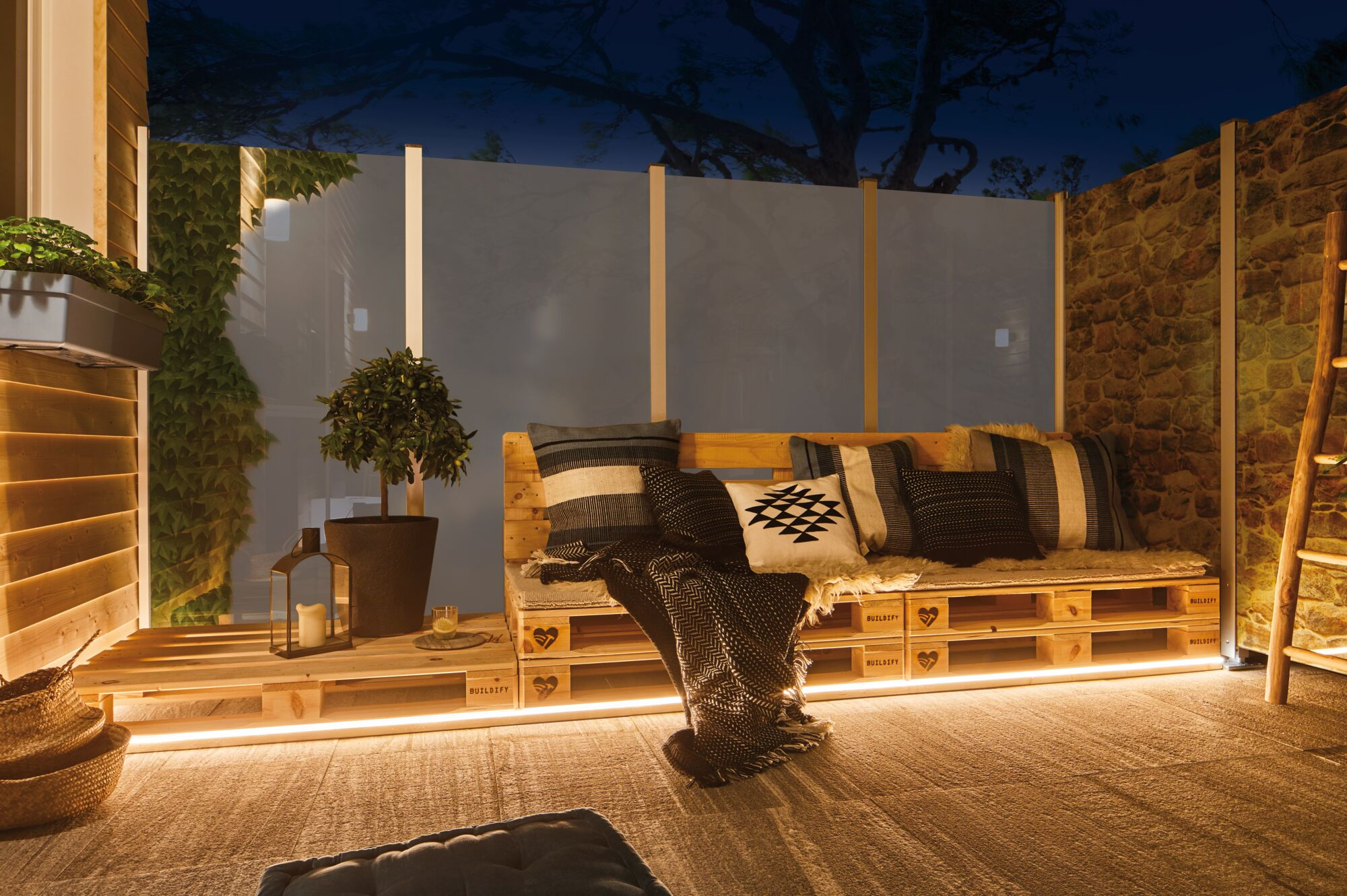 Beleuchtetes Europaletten-Sofa auf einem Balkon mit Kissen, Topfpflanze und warmer Bodenbeleuchtung.
