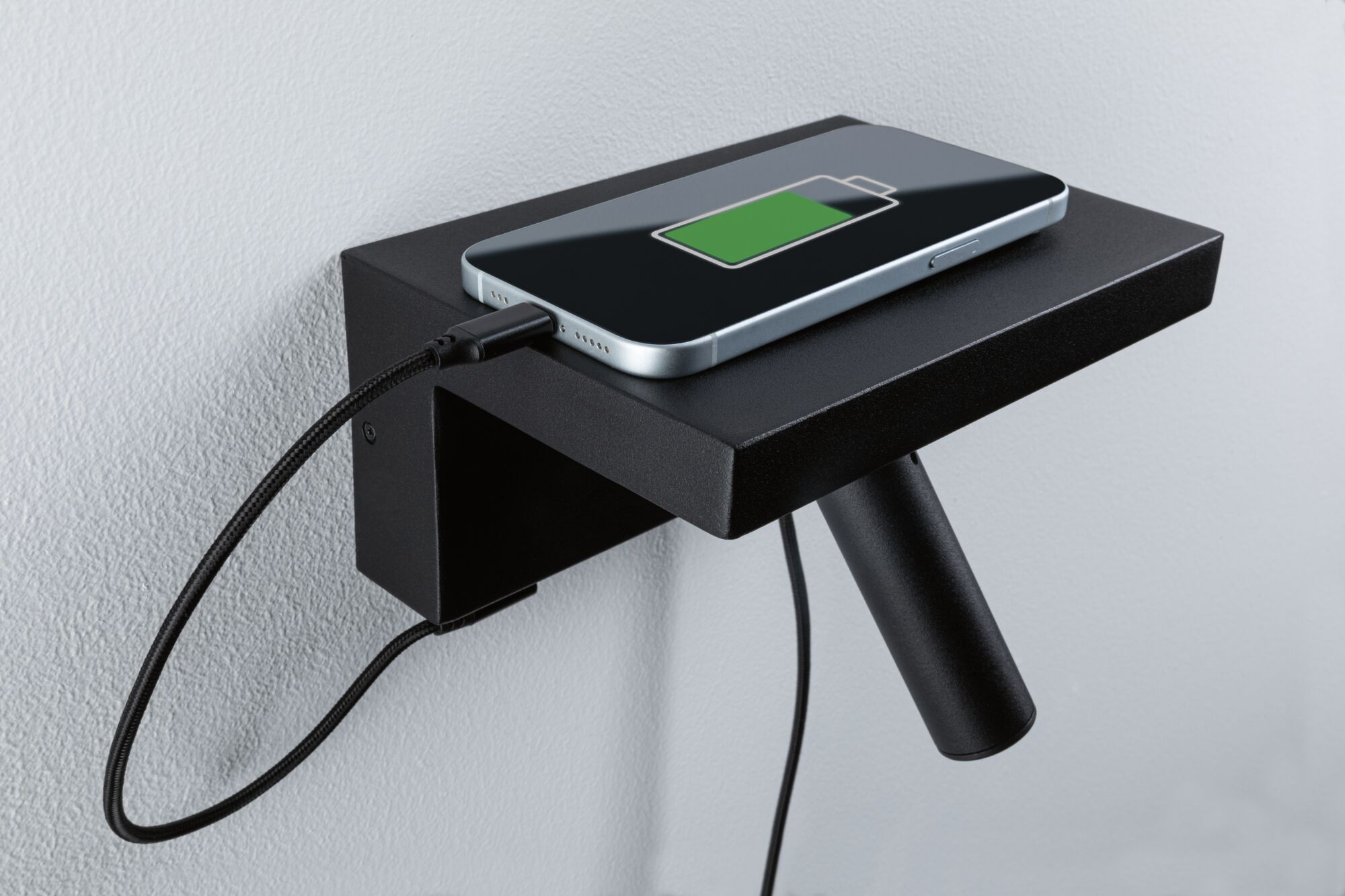 Ein Smartphone lädt auf einem kleinen schwarzen Wandregal gegen eine helle Wand.