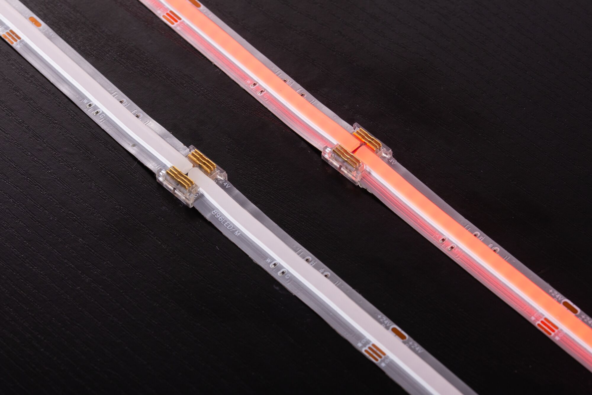 Zwei LED-Leuchtstreifen mit Steckverbindern liegen auf einer dunklen Holzoberfläche, einer leuchtet weiß, der andere orange.
