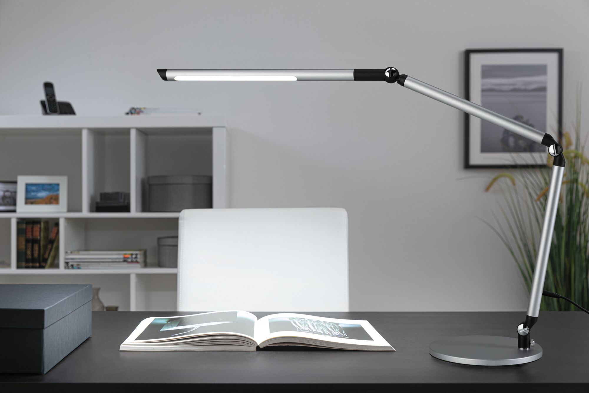 Eine silberne moderne Schreibtischlampe leuchtet auf einem dunklen Tisch mit offenem Buch.