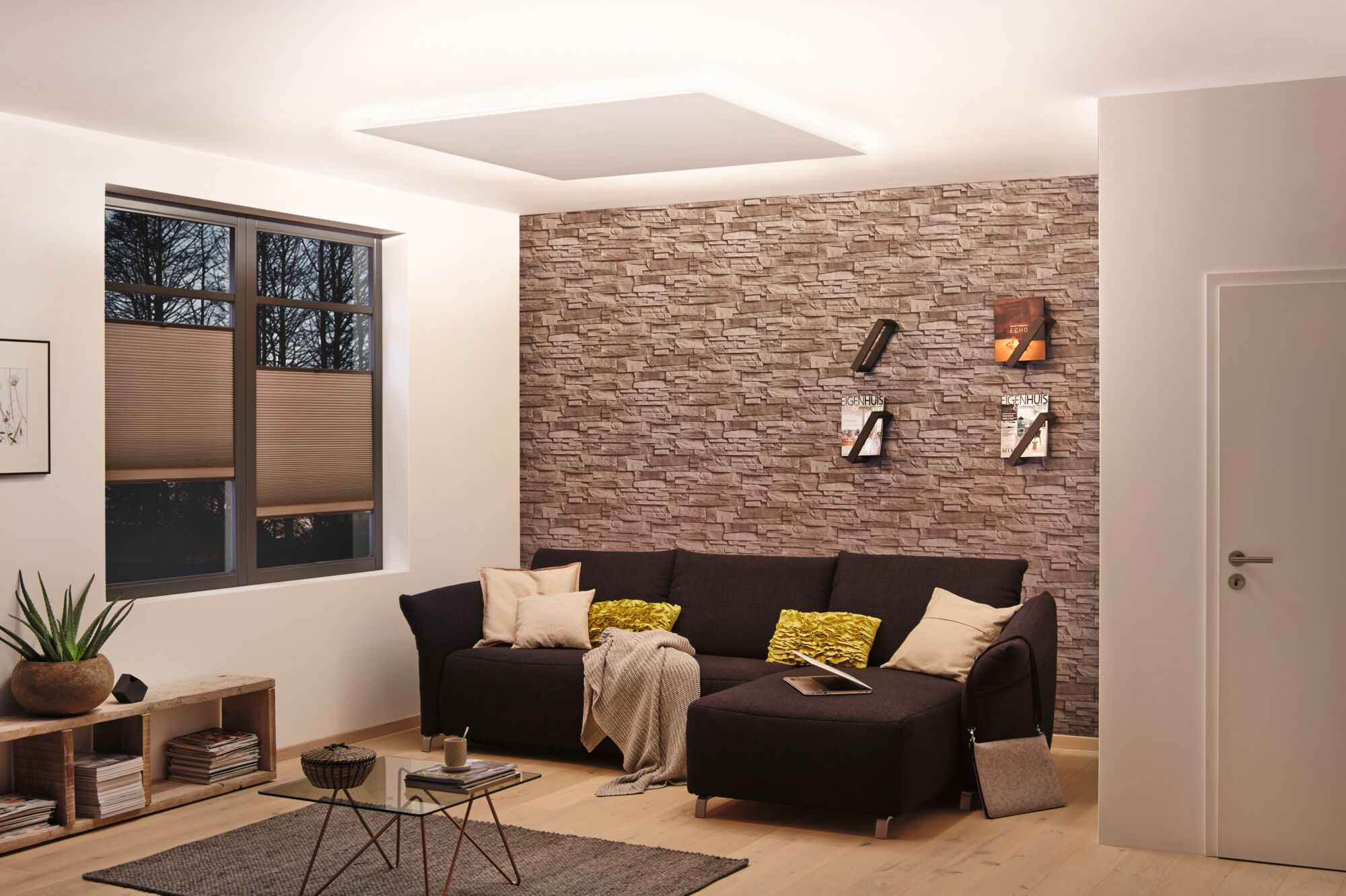 Moderne Wohnzimmer mit braunem Sofa, Natursteinwand, Glastisch und großem Fenster mit Plissee.
