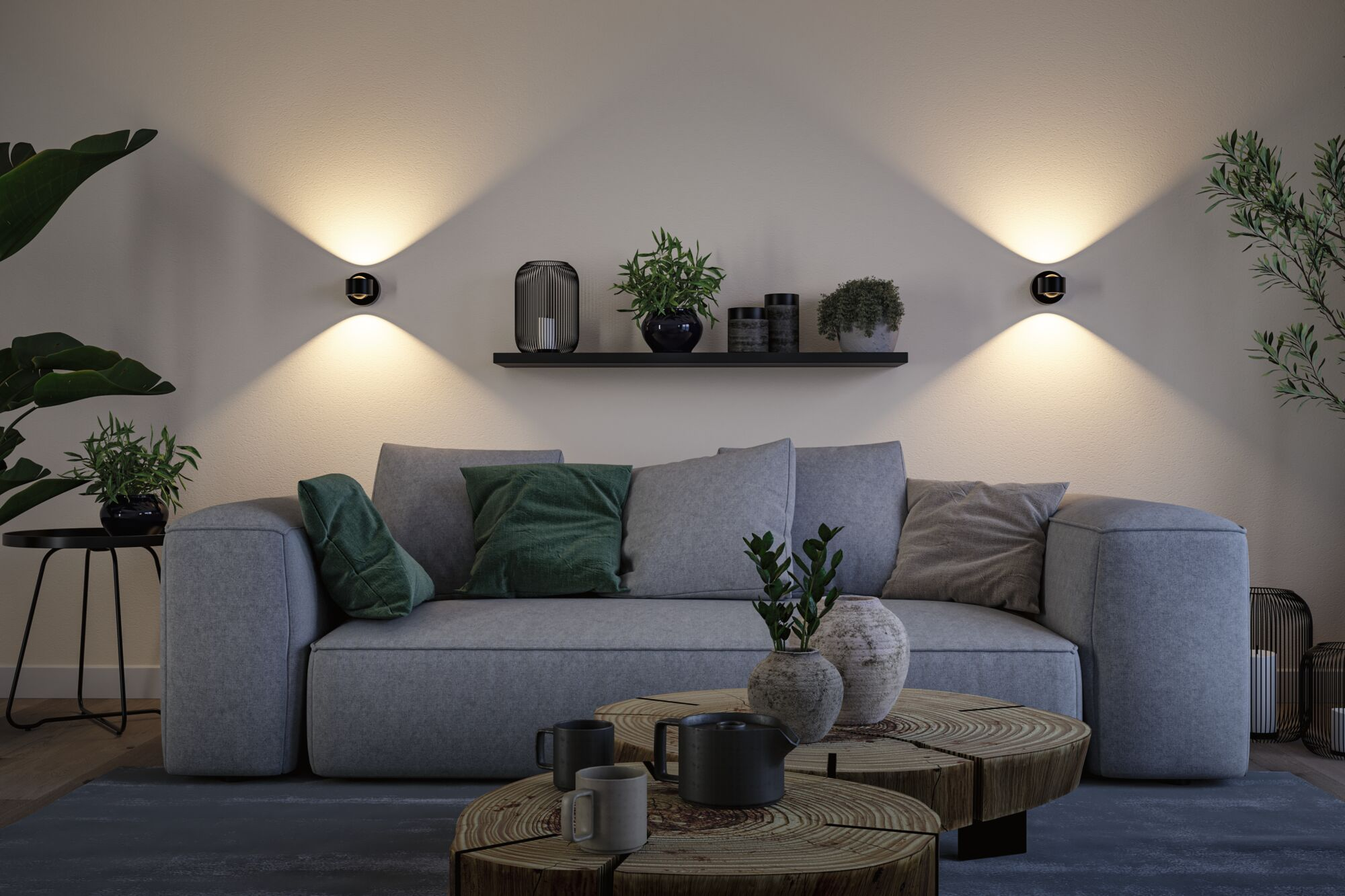 Ein graues Sofa mit grünen Kissen, Holztischen, Pflanzen und zwei Wandleuchten im warmen Licht.