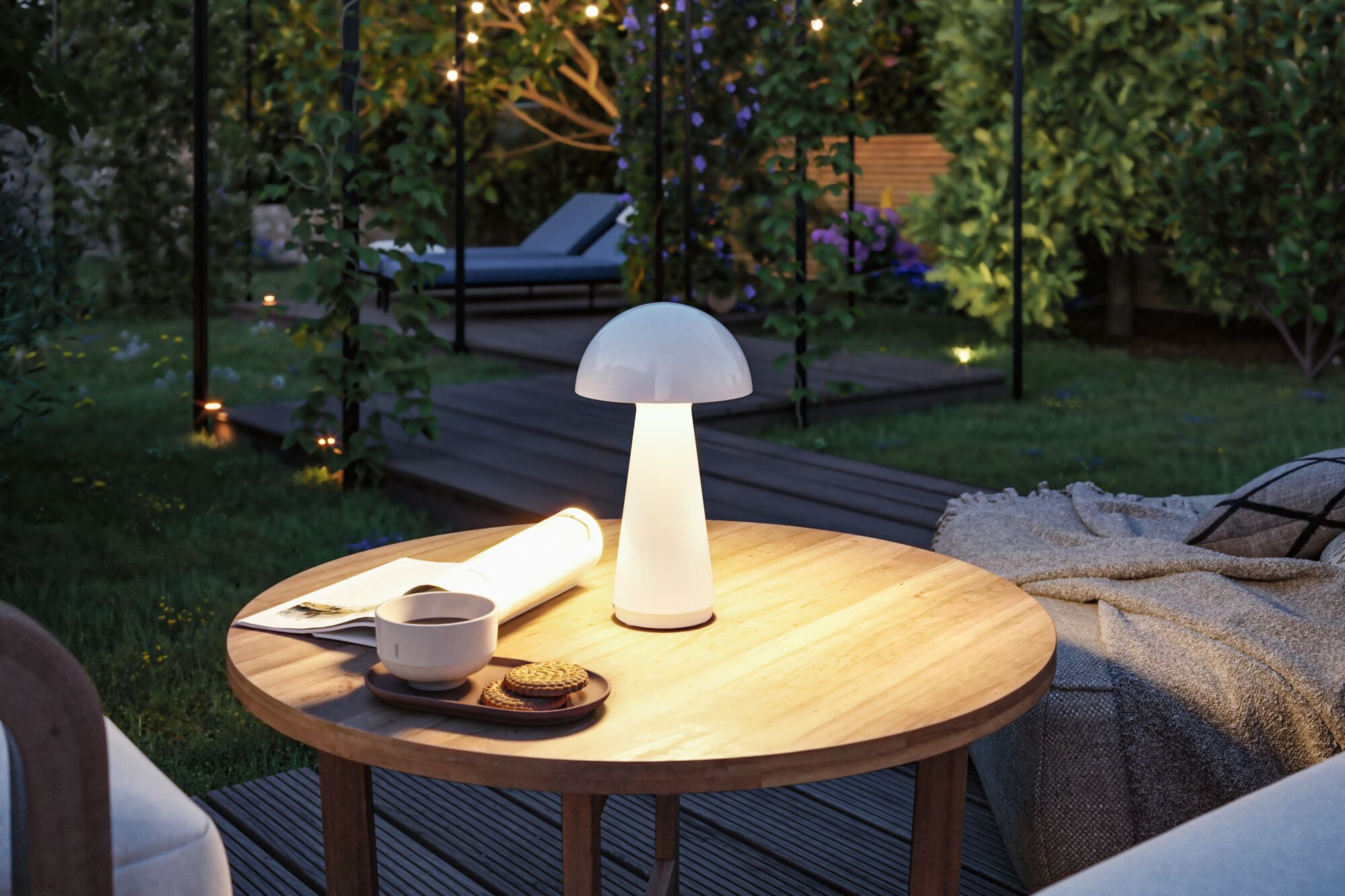 Eine weiße Tischlampe, ein Buch, eine Kaffeetasse und Kekse auf einem runden Holztisch im Garten bei Nacht.