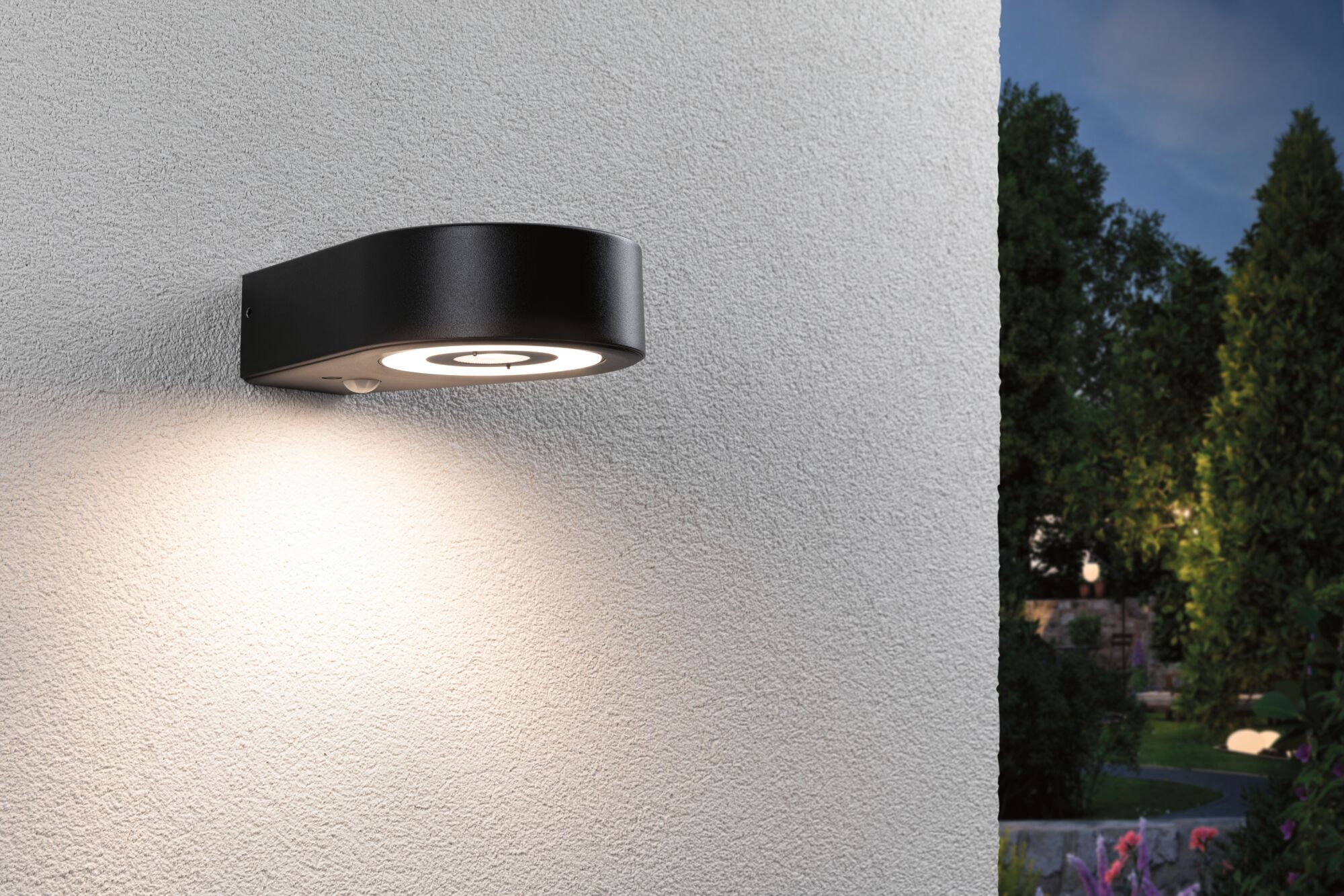 Eine schwarze Außenwandleuchte mit integriertem Licht strahlt an einer weißen verputzten Wand.