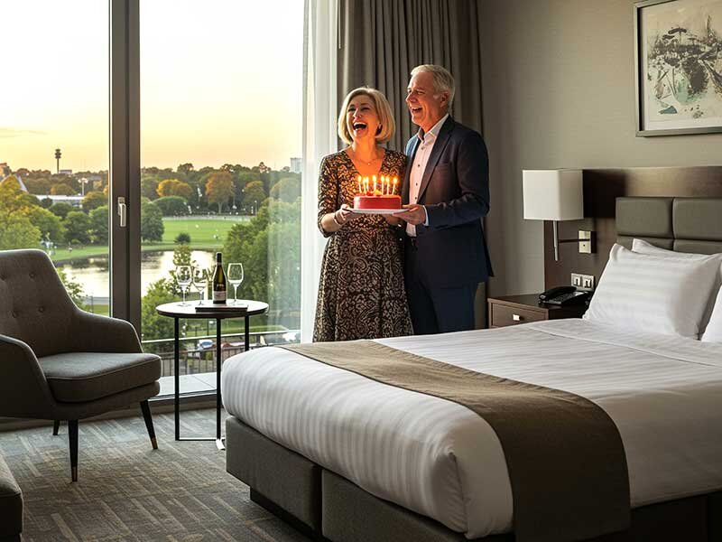 Una pareja feliz sostiene un pastel de cumpleaños con velas en una habitación de hotel con vista al parque.