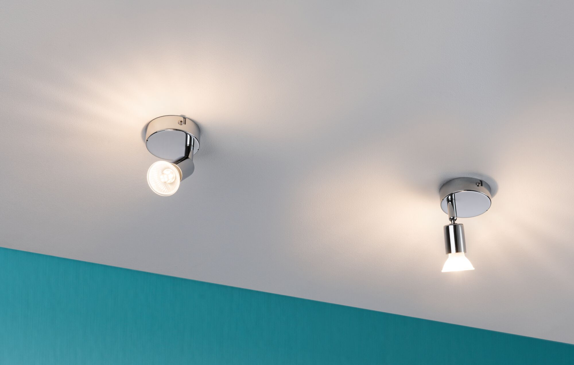 Zwei verchromte Strahler an einer weißen Decke über einer türkisfarbenen Wand, beide Lampen sind eingeschaltet.