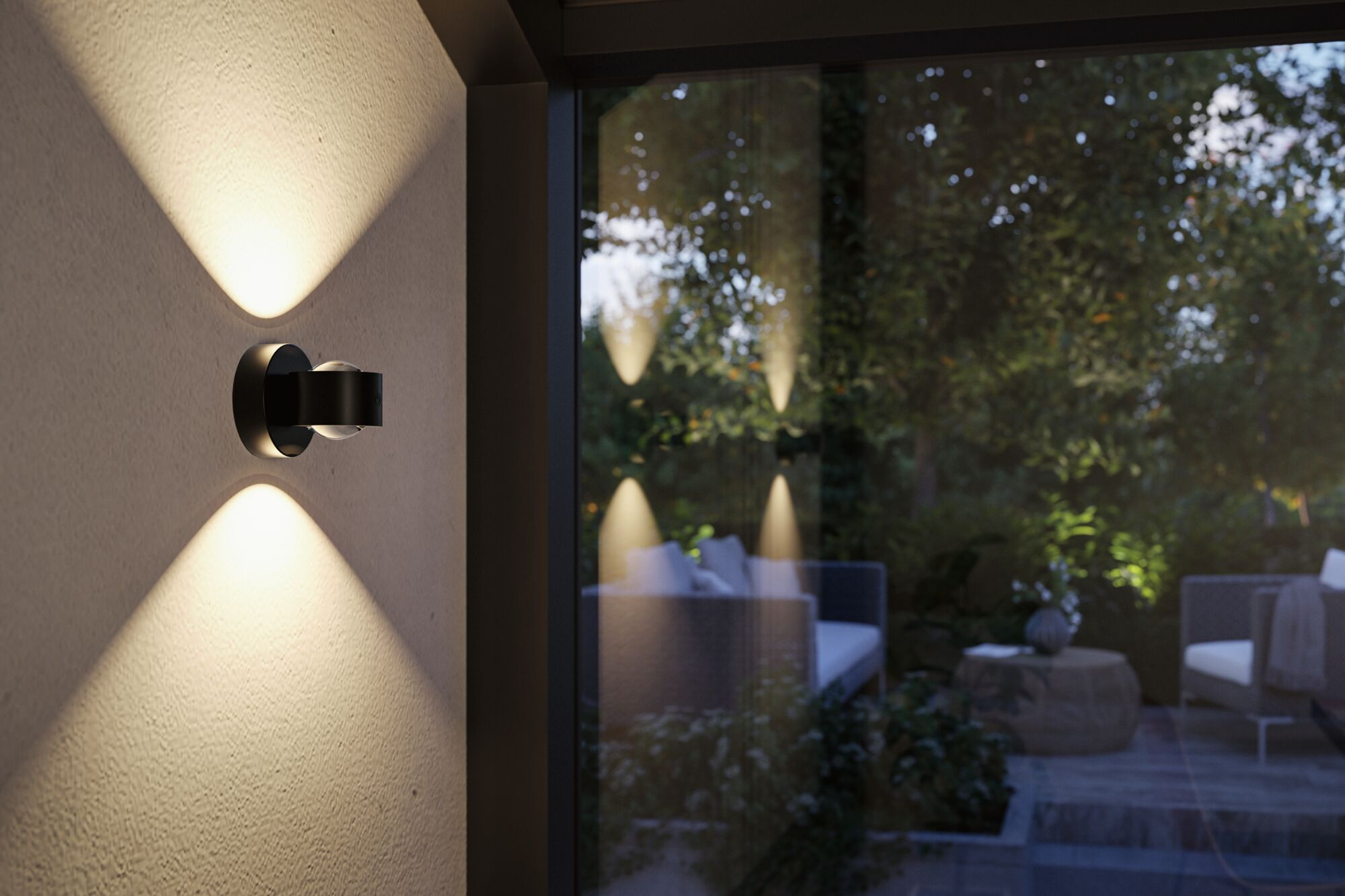 Eine schwarze Wandlampe strahlt an einer Wand nach oben und unten, im Hintergrund ist ein Garten zu sehen.