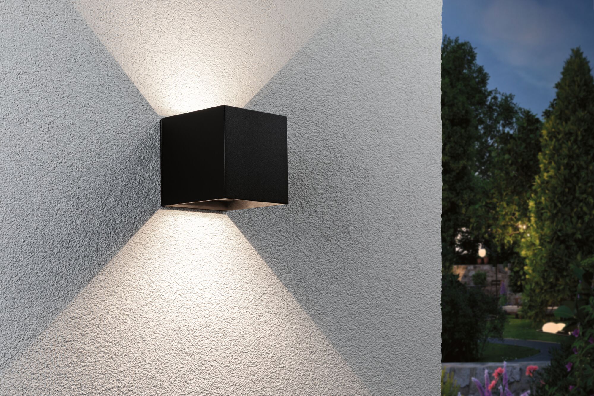 Eine schwarze, würfelförmige Wandleuchte an einer strukturierten Außenwand wirft in der Dämmerung Lichtkegel.