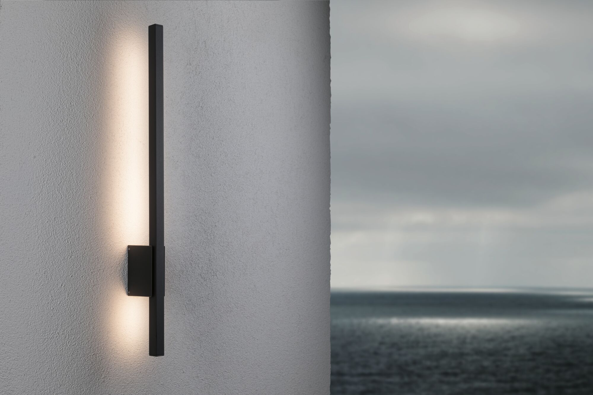 Schlanke schwarze Wandleuchte an einer grauen Wand mit Blick auf das Meer im Hintergrund.