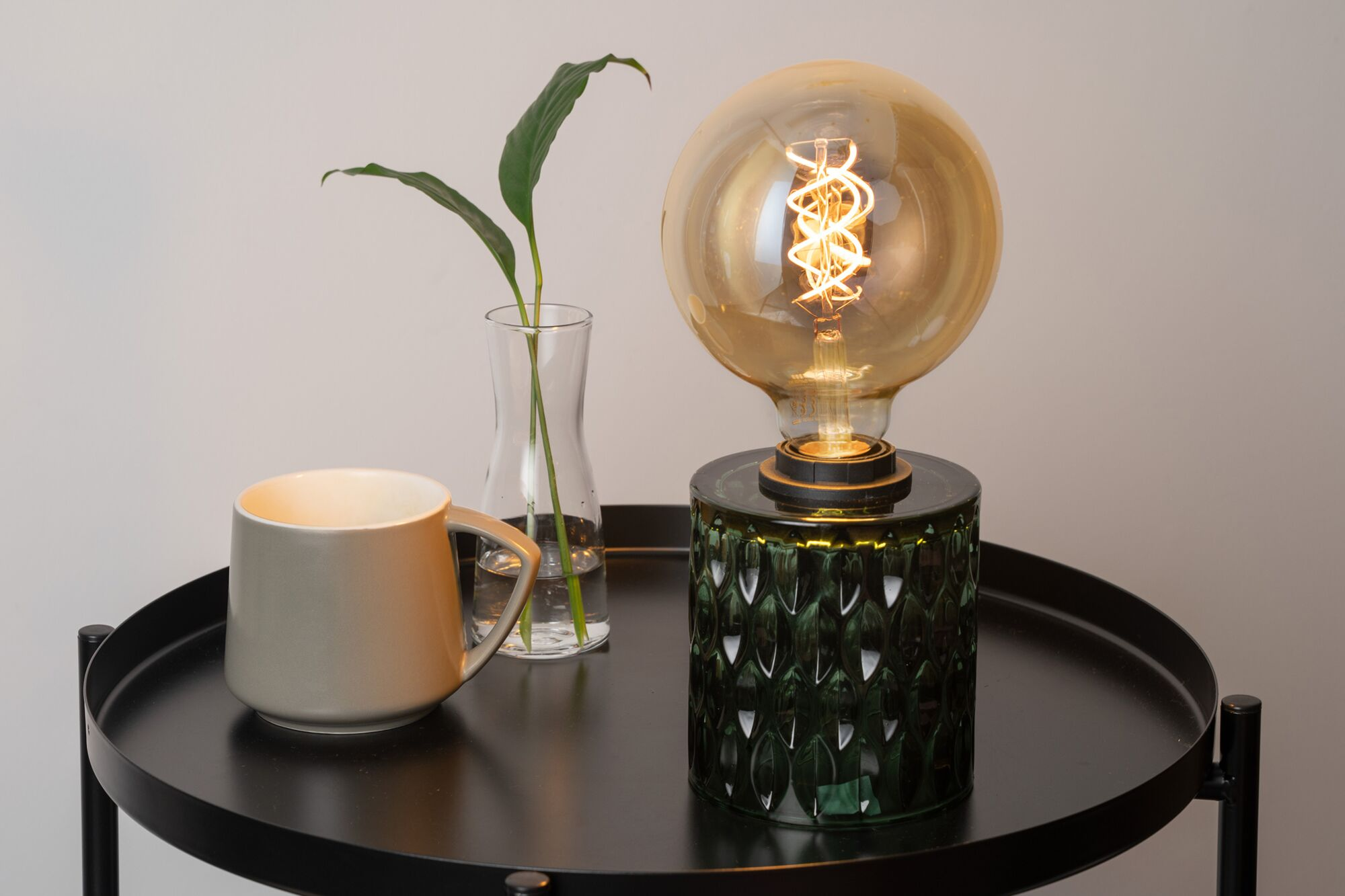 Eine brennende Edison-Lampe auf einem strukturierten grünen Glasfuss, neben einer Glasvase und einer grauen Tasse.