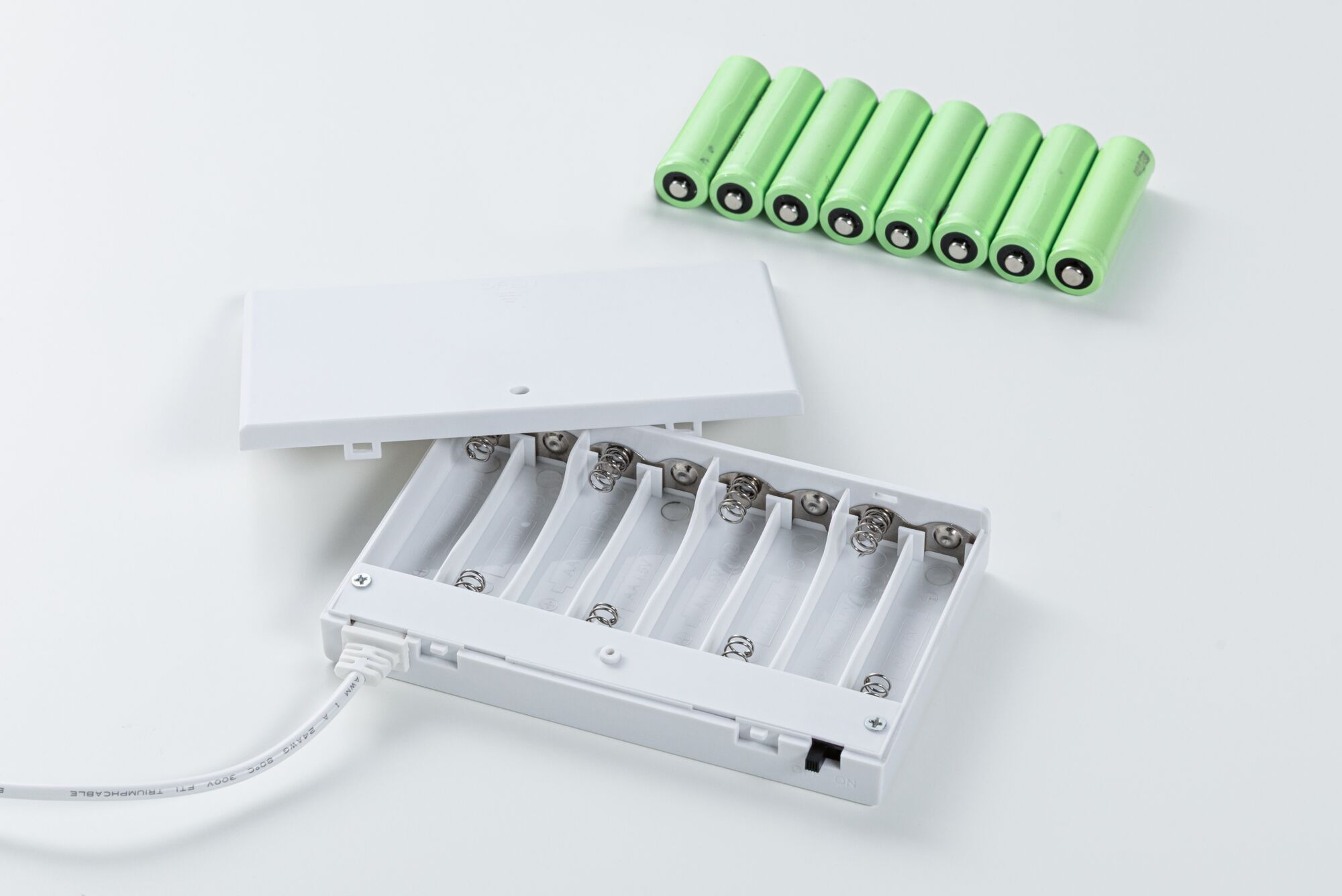 Ein weißes Batteriegehäuse mit offener Abdeckung und ein Kabel, daneben acht grüne AA-Akkus.
