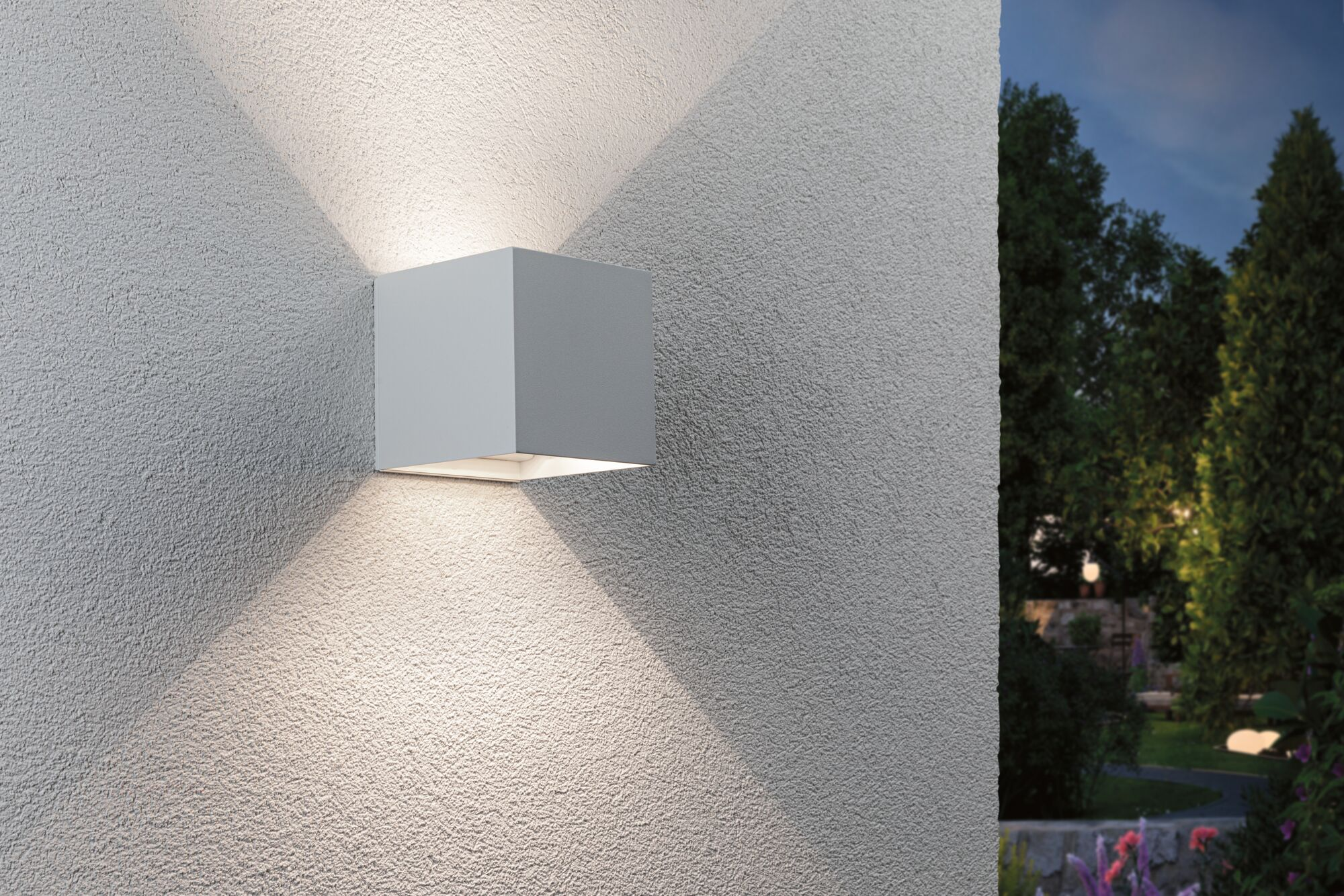 Eine moderne quadratische Wandleuchte wirft Lichtkegel an eine strukturierte Außenwand mit Gartenblick.