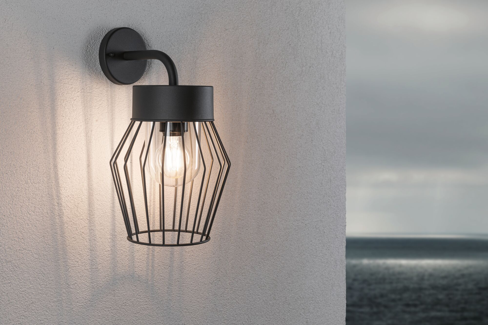 Eine schwarze Wandlampe mit Metallgitter und sichtbarer Glühbirne an einer hellen Wand, im Hintergrund das Meer.