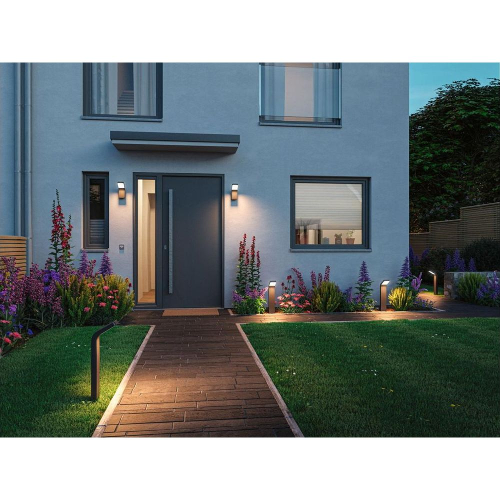 Beleuchteter Gehweg führt zu einer grauen Haustür eines modernen Hauses, umgeben von blühenden Pflanzen und Gartenleuch