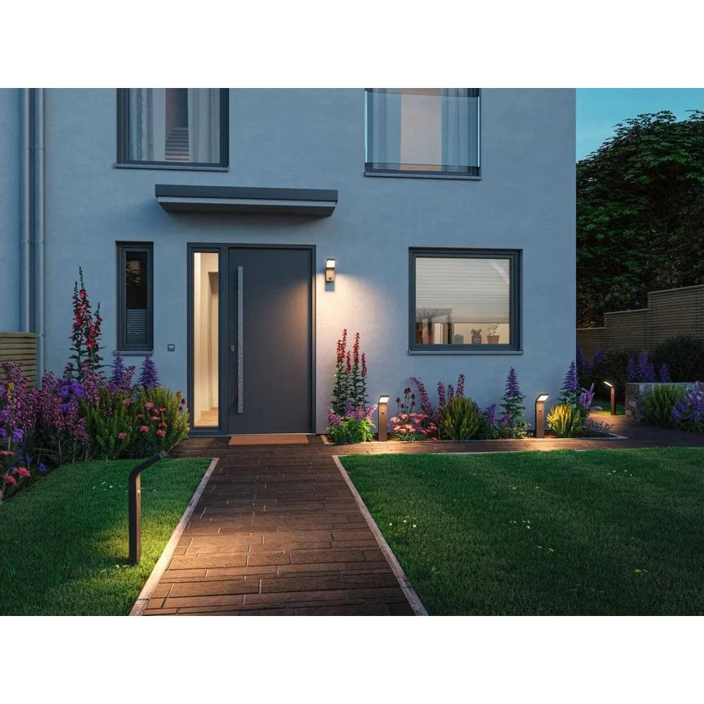 Beleuchteter Gehweg führt zu einer grauen Haustür eines modernen Hauses, umgeben von blühendem Garten bei Dämmerung.