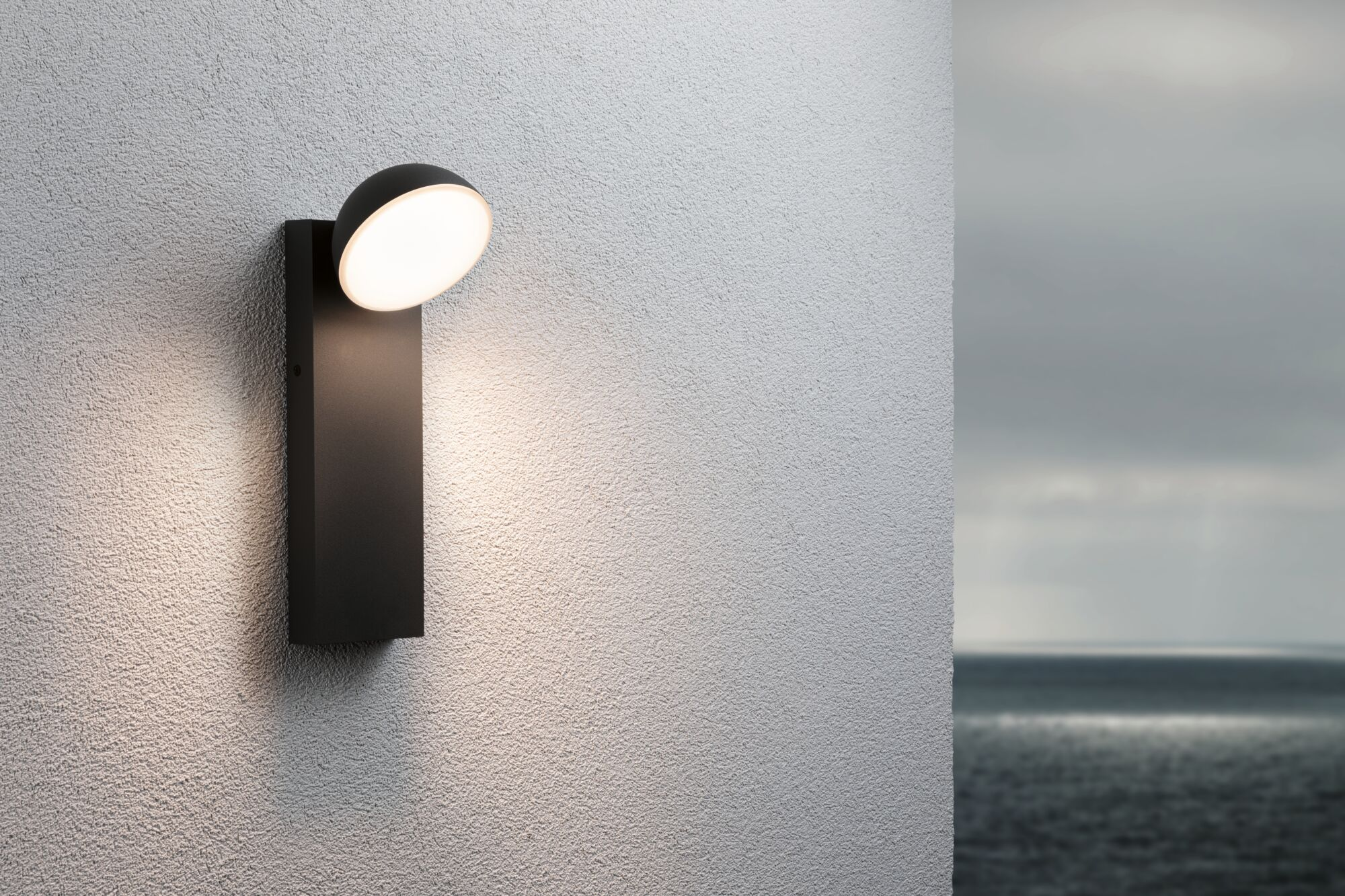 Eine moderne schwarze Außenwandleuchte leuchtet an einer strukturierten weißen Wand am Meer.