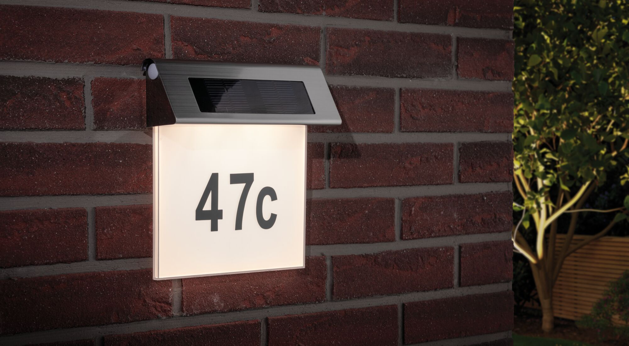 Ein leuchtendes Hausschild mit der Nummer 47c an einer roten Ziegelwand mit Solarpanel.