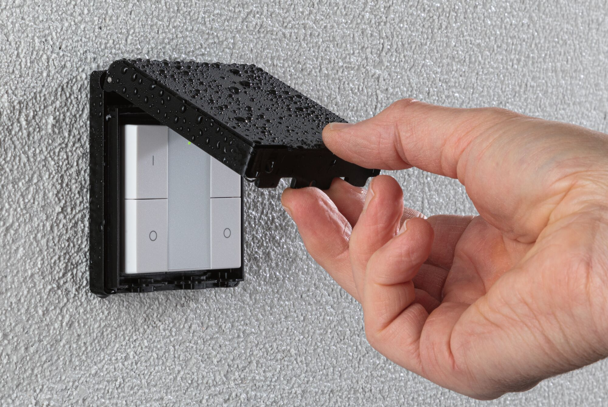 Eine Hand öffnet die feuchte Schutzabdeckung eines weißen Lichtschalters an einer grauen Wand.