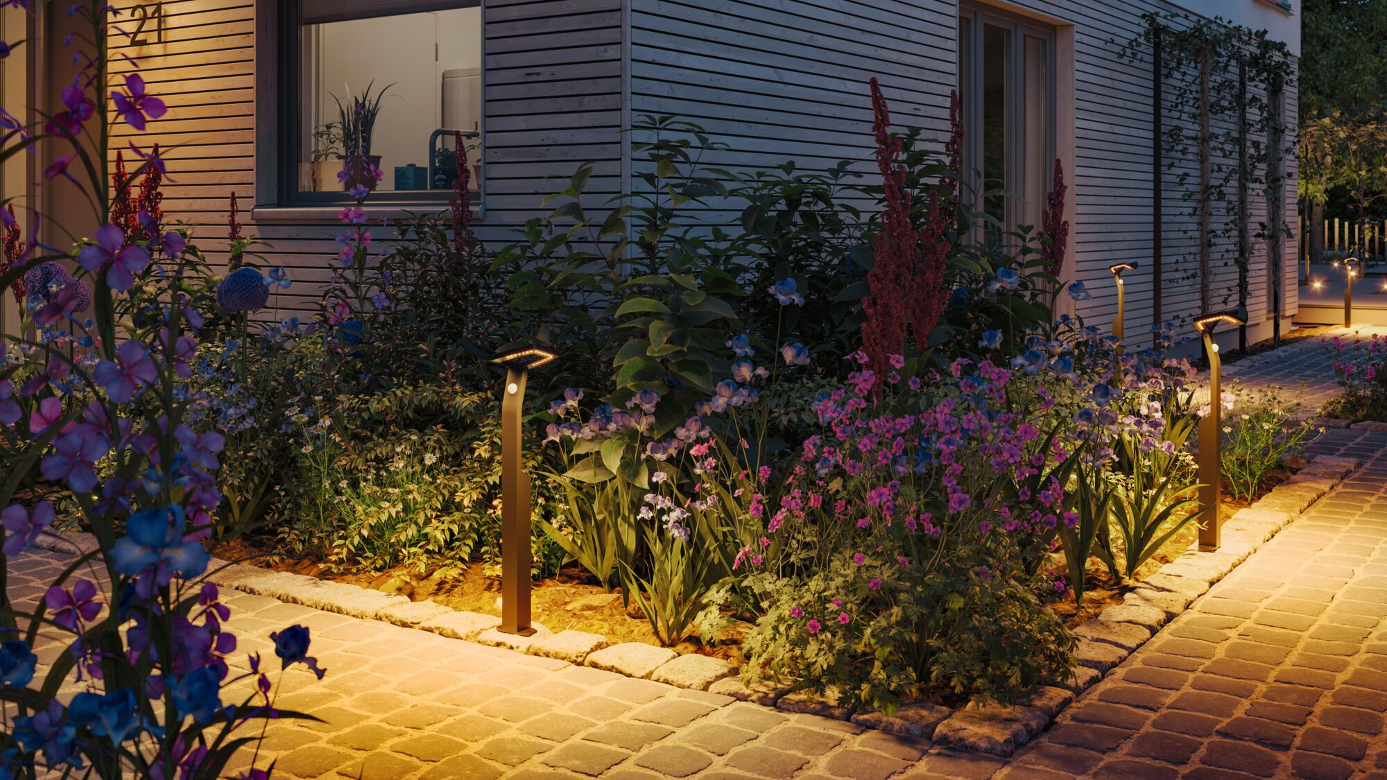 Beleuchteter Gartenweg im Dunkeln mit modernen Wegeleuchten, bunten Blumenbeeten und einer hellen Hausfassade.