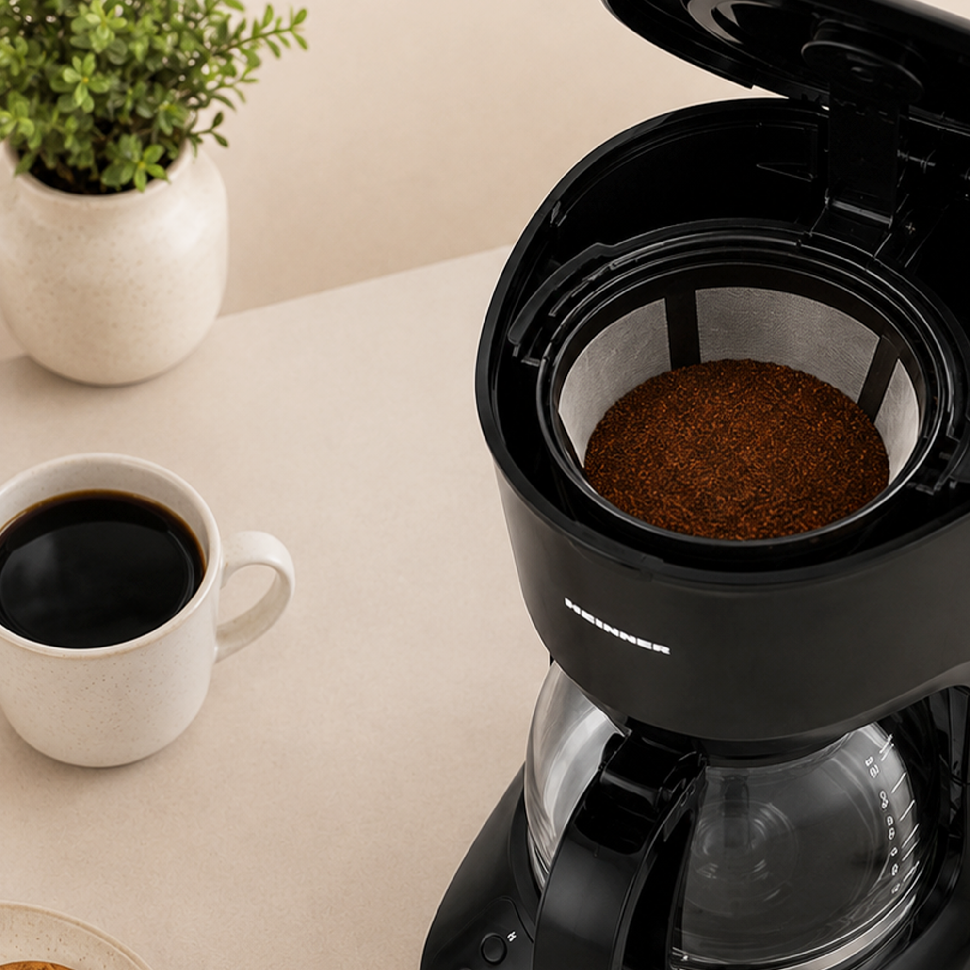 Eine schwarze Filterkaffeemaschine mit Kaffeemehl, eine weiße Tasse Schwarzer Kaffee und eine Topfpflanze.