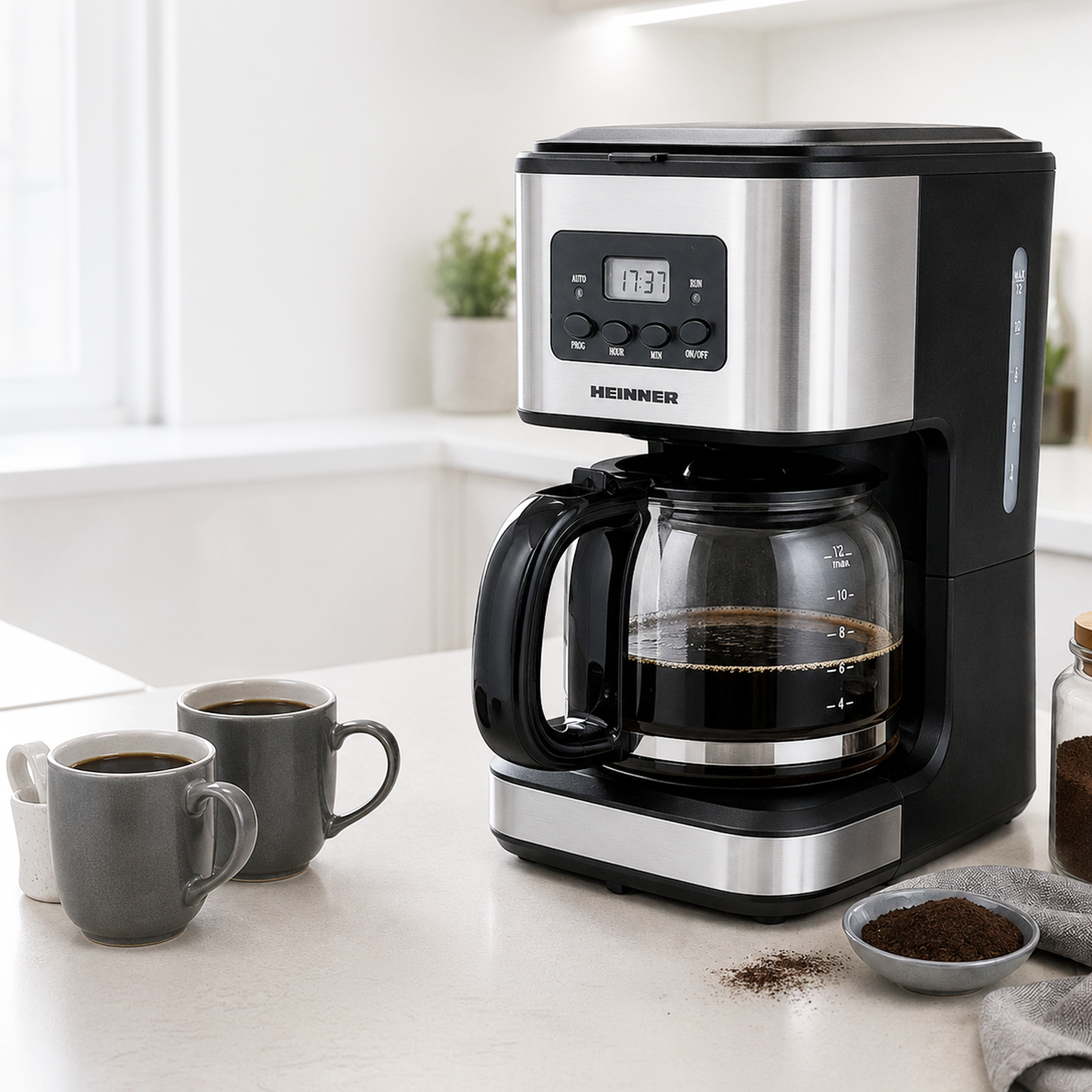 A black and silver Heinner coffee maker sits on a kitchen counter beside two grey mugs.